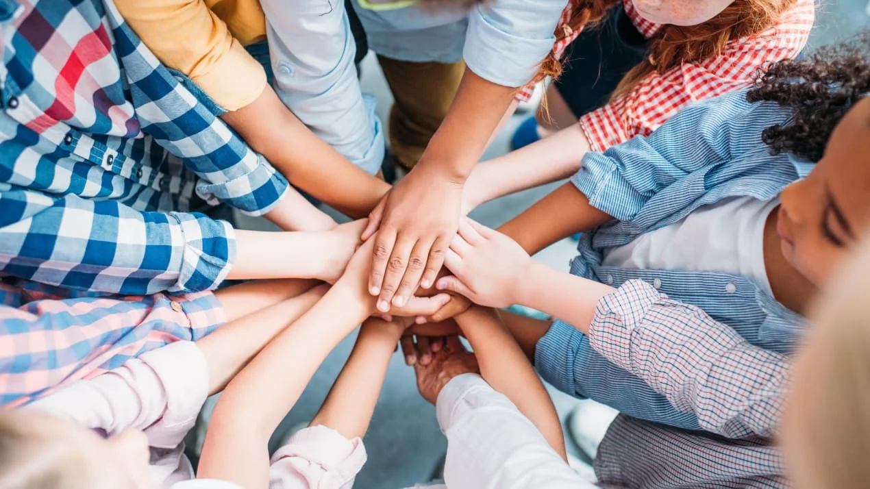 A group of children with different skin tones stacking their hands together in the middle, symbolizing teamwork and unity.