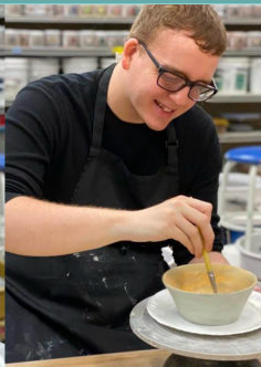 A young man with glasses smiling while stirring a bowl of batter or mixture in a kitchen or food prep area.