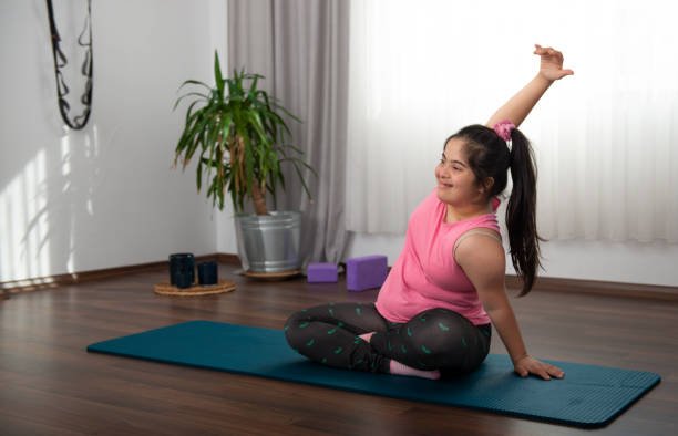 Young girl in pink workout top and leggings sitting cross-legged on yoga mat, stretching arm over head in a bright room with wooden floor, large window with curtains, potted plant, and exercise equipment in background.