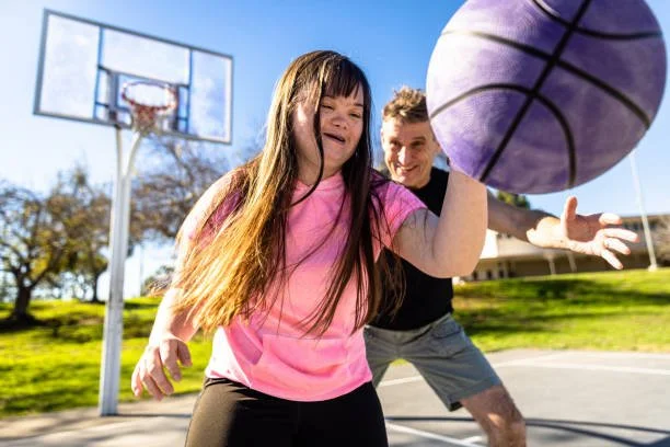 A girl in a pink shirt and a man outside on a basketball court, with a basketball in the foreground. The girl appears to be reaching for the ball and the man is smiling in the background.