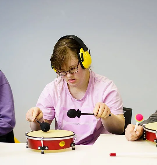 A young girl in glasses and pink shirt wearing yellow noise-canceling headphones playing a toy drum with mallets.