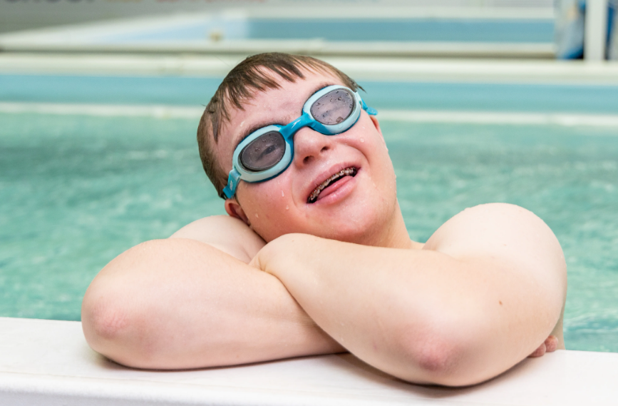 A young boy wearing goggles leaning on the edge of a swimming pool with water in the background.