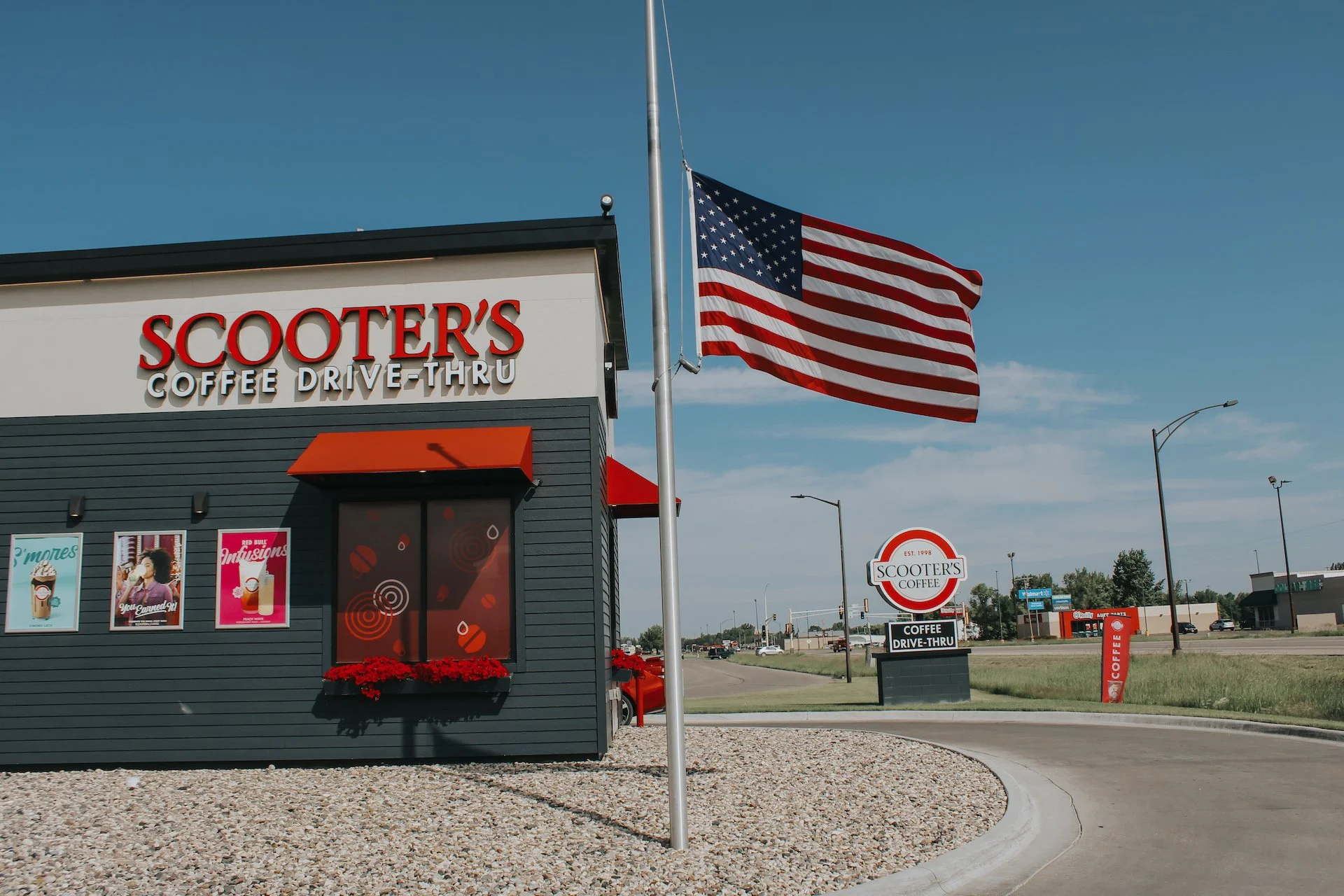 Exterior of Scooter's Coffee with a large American flag on a flagpole, a sign indicating drive-thru service, and advertisements on the building under a clear blue sky.