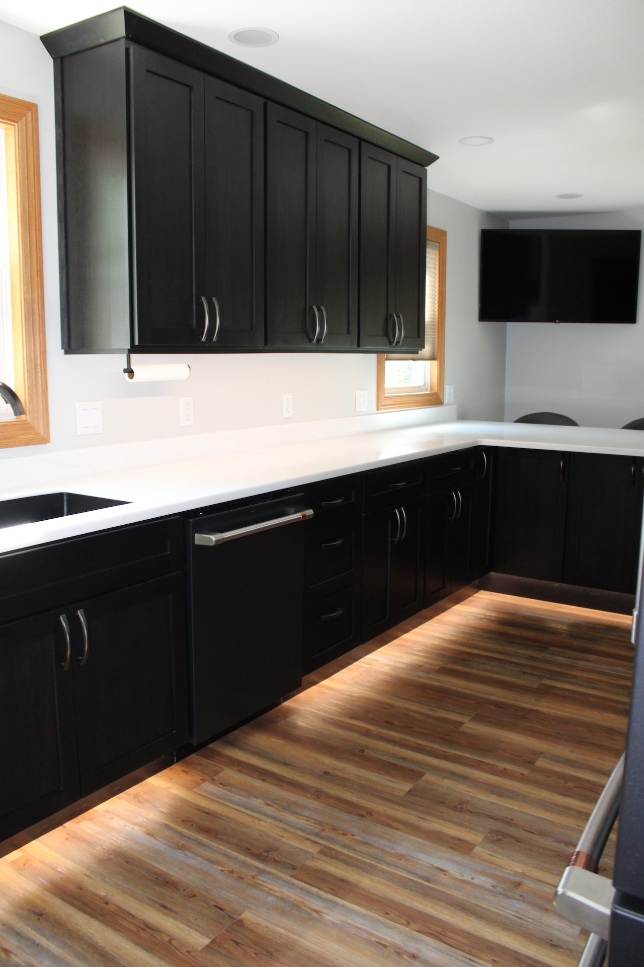 Kitchen with black cabinets, white countertops, wooden floors, windows, and a wall-mounted flat-screen TV