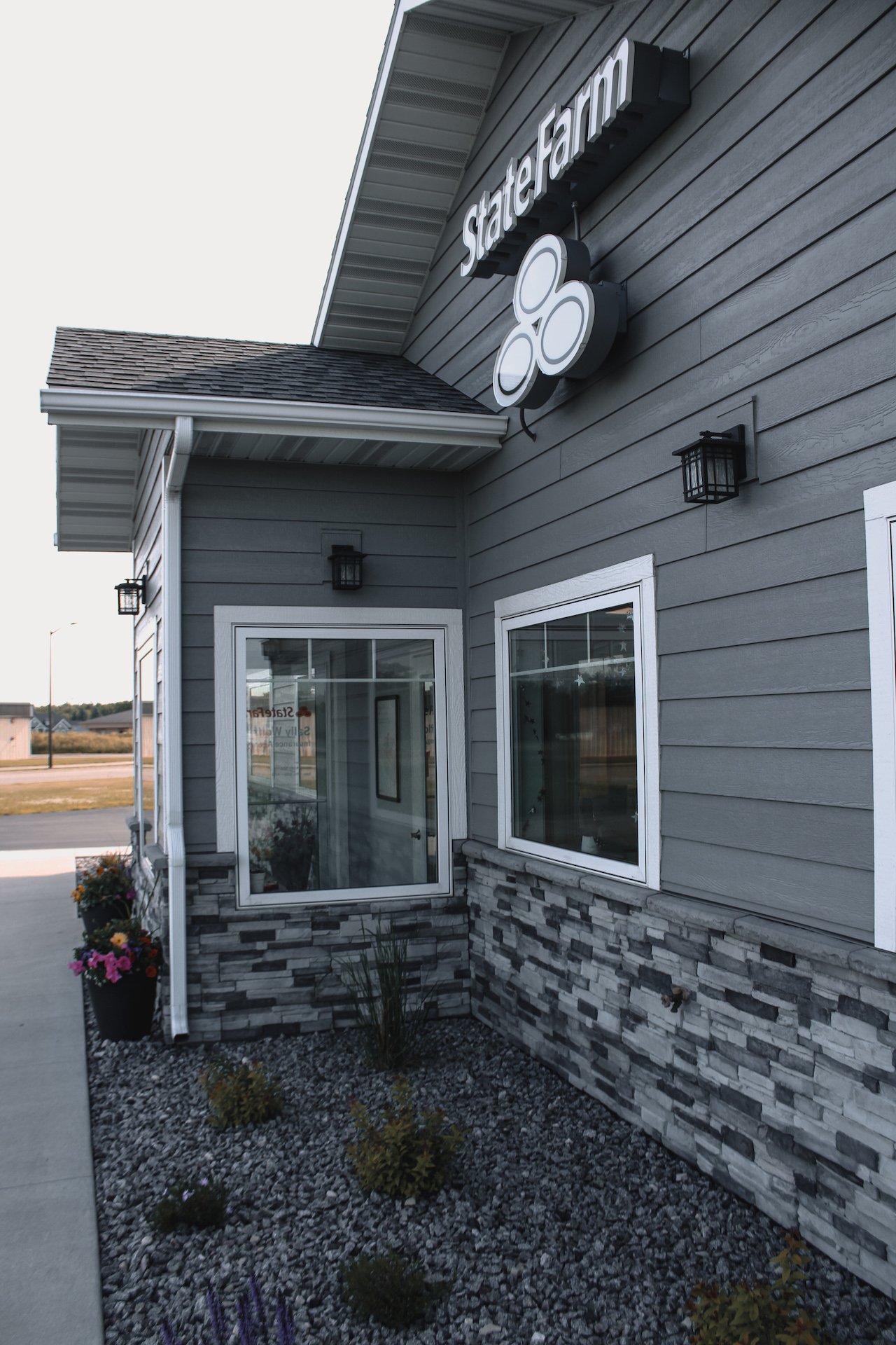 Exterior of a State Farm insurance office with gray siding, white trim, and a sign featuring the State Farm logo with three overlapping ovals.