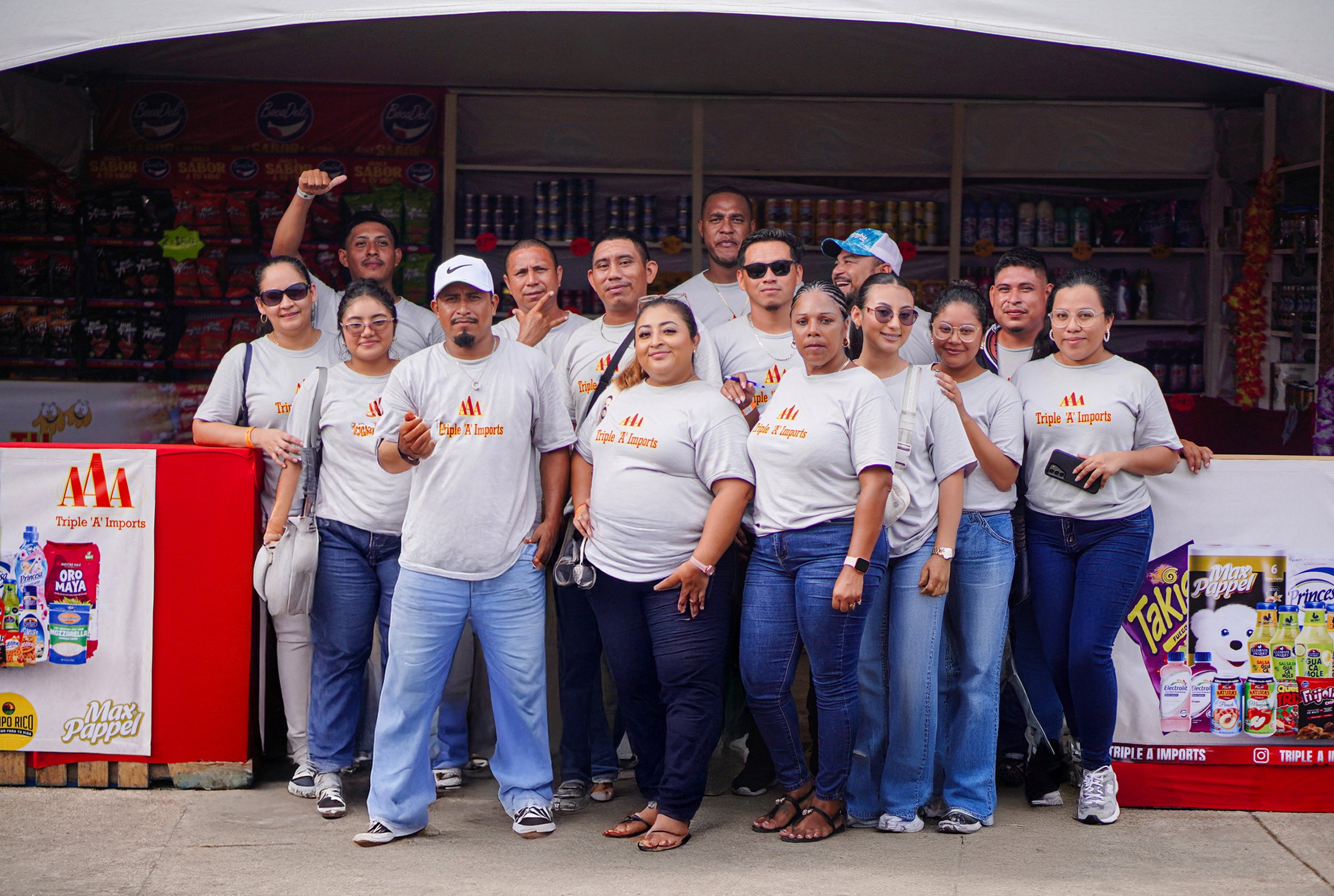 A group of people wearing white T-shirts with a logo, standing in front of a small store or booth that sells snacks and household items. They are posing for a photo, some smiling and some making gestures.
