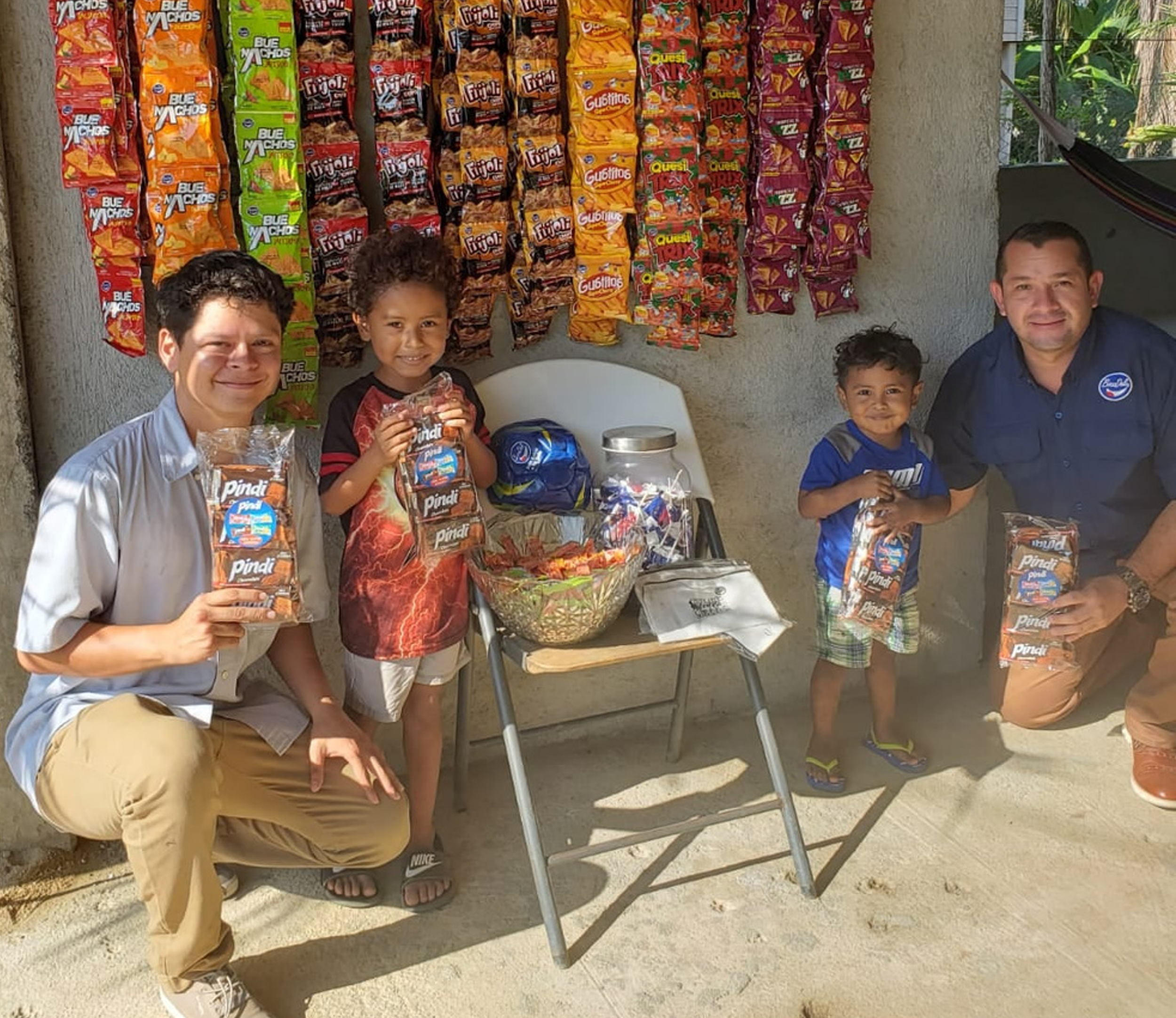 A group of four smiling people, two adults and two children, holding bags of Pindi snacks, standing outdoors in front of a wall decorated with colorful snack packages. There is a small table with a bowl of snacks and additional items.