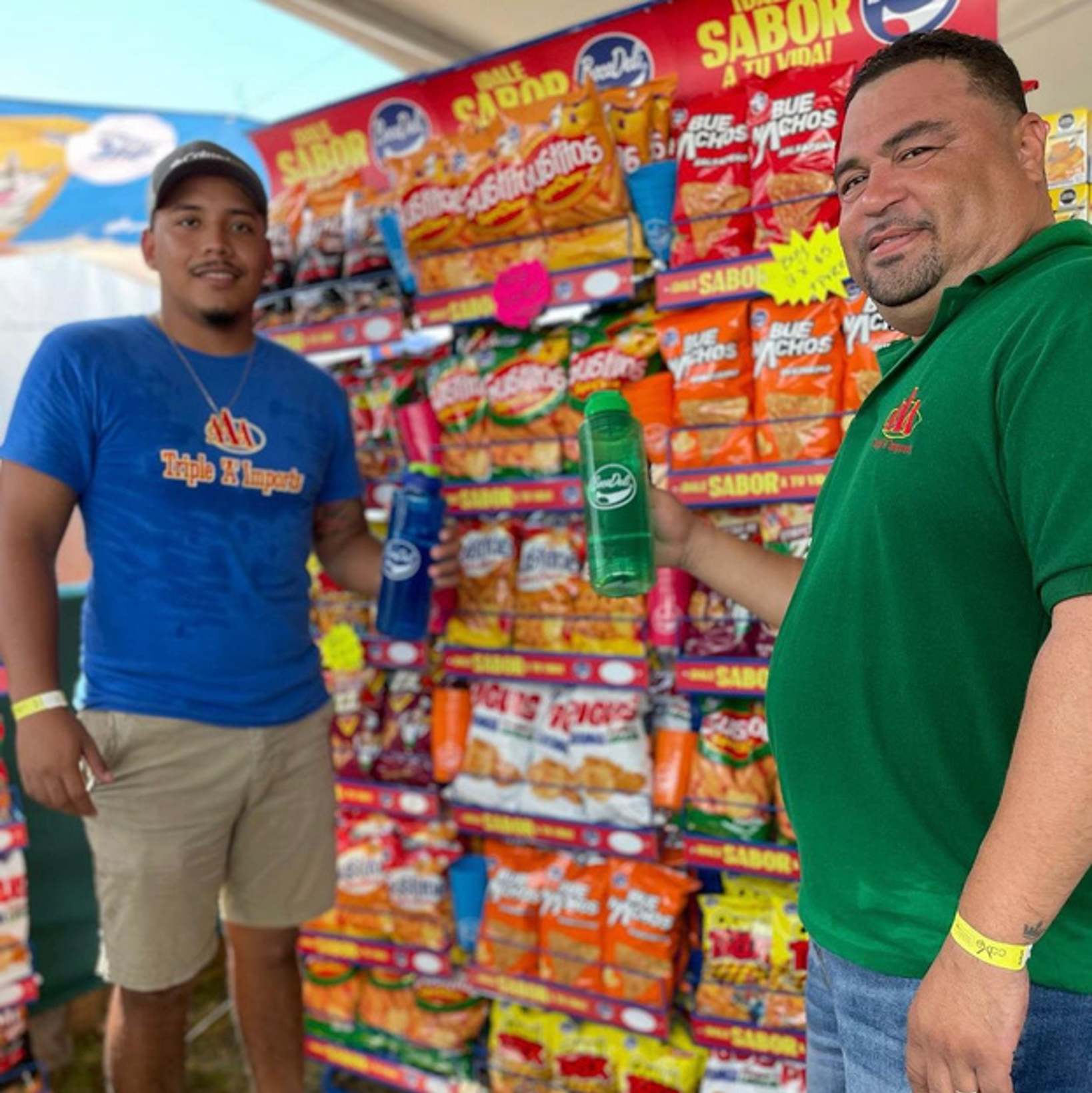 Two men standing in front of a display of snack bags at a fair or market. The man on the left wears a blue t-shirt and beige shorts, holding a blue bottle. The man on the right wears a green polo shirt and jeans, holding a green bottle. The snack display has red, orange, and yellow packaging.