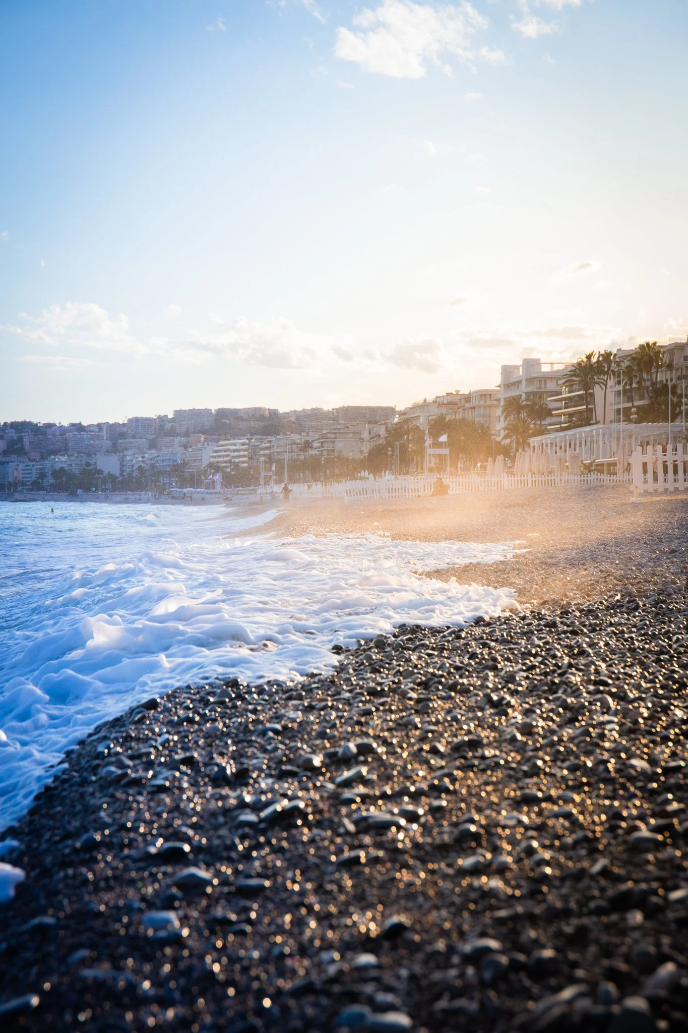 Pebble beach with waves and seafront buildings at sunset in Nice, France.