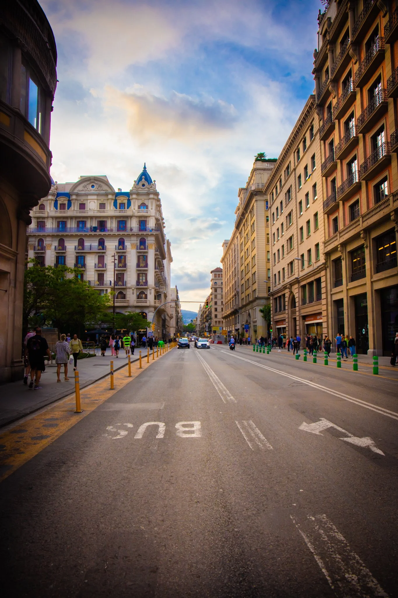 Street scene in a European city with historic buildings, pedestrians, and cars. Sidewalks are lined with bollards, and the sky is partly cloudy.