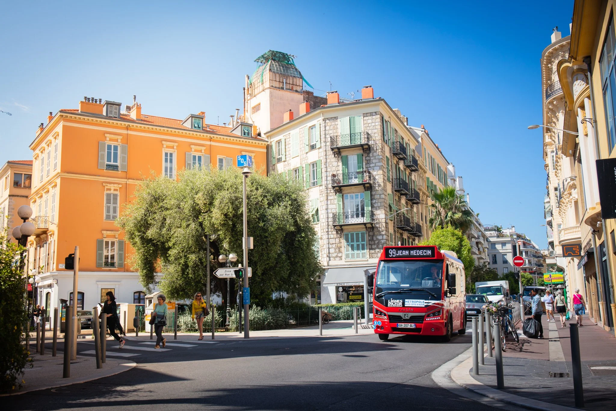 Street in a European city with a red bus labeled '99 Jean Medecin,' people walking, and colorful buildings with balconies and greenery.