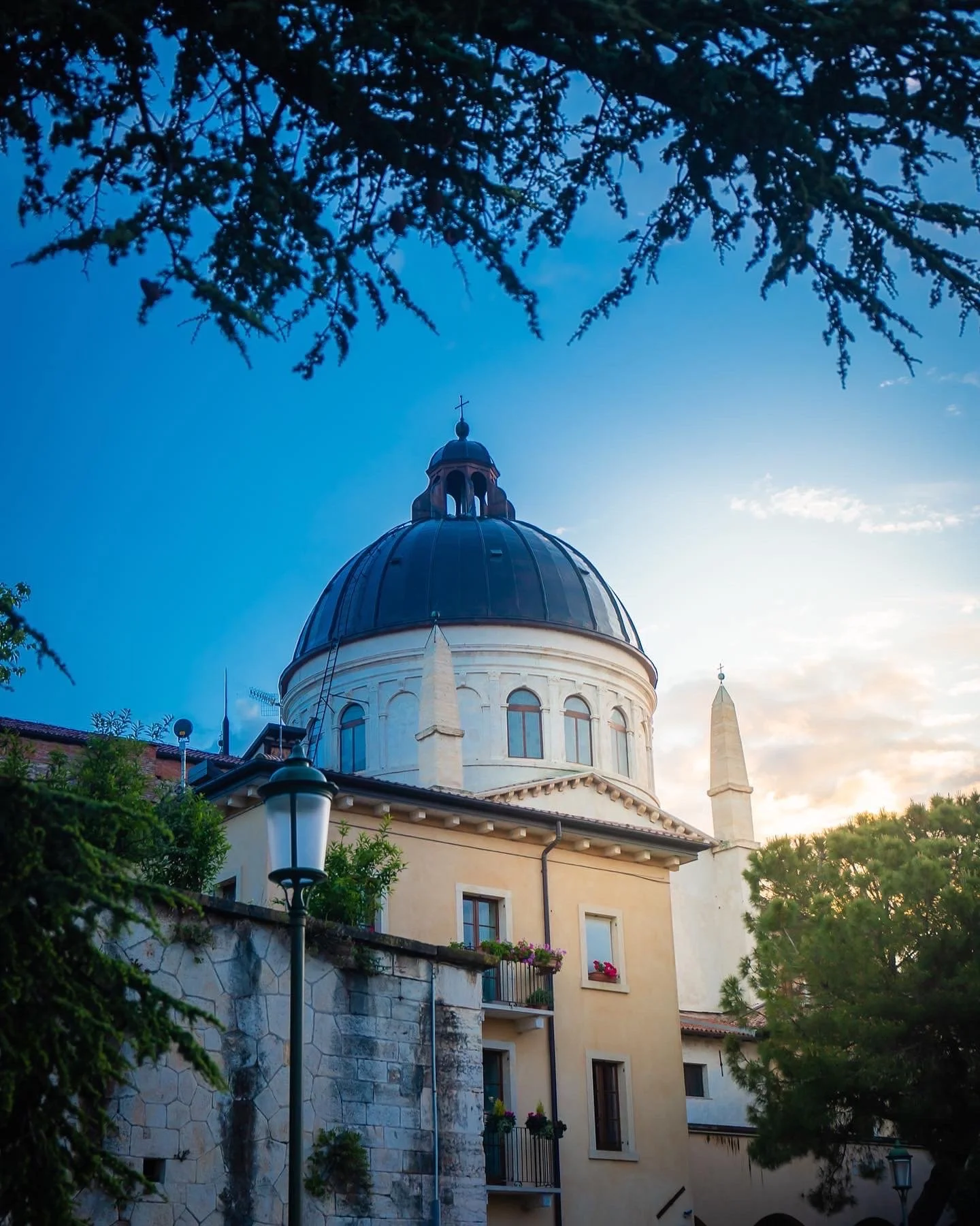 Historical building with a dome and spire, surrounded by trees and lamppost, under a blue sky at sunset.