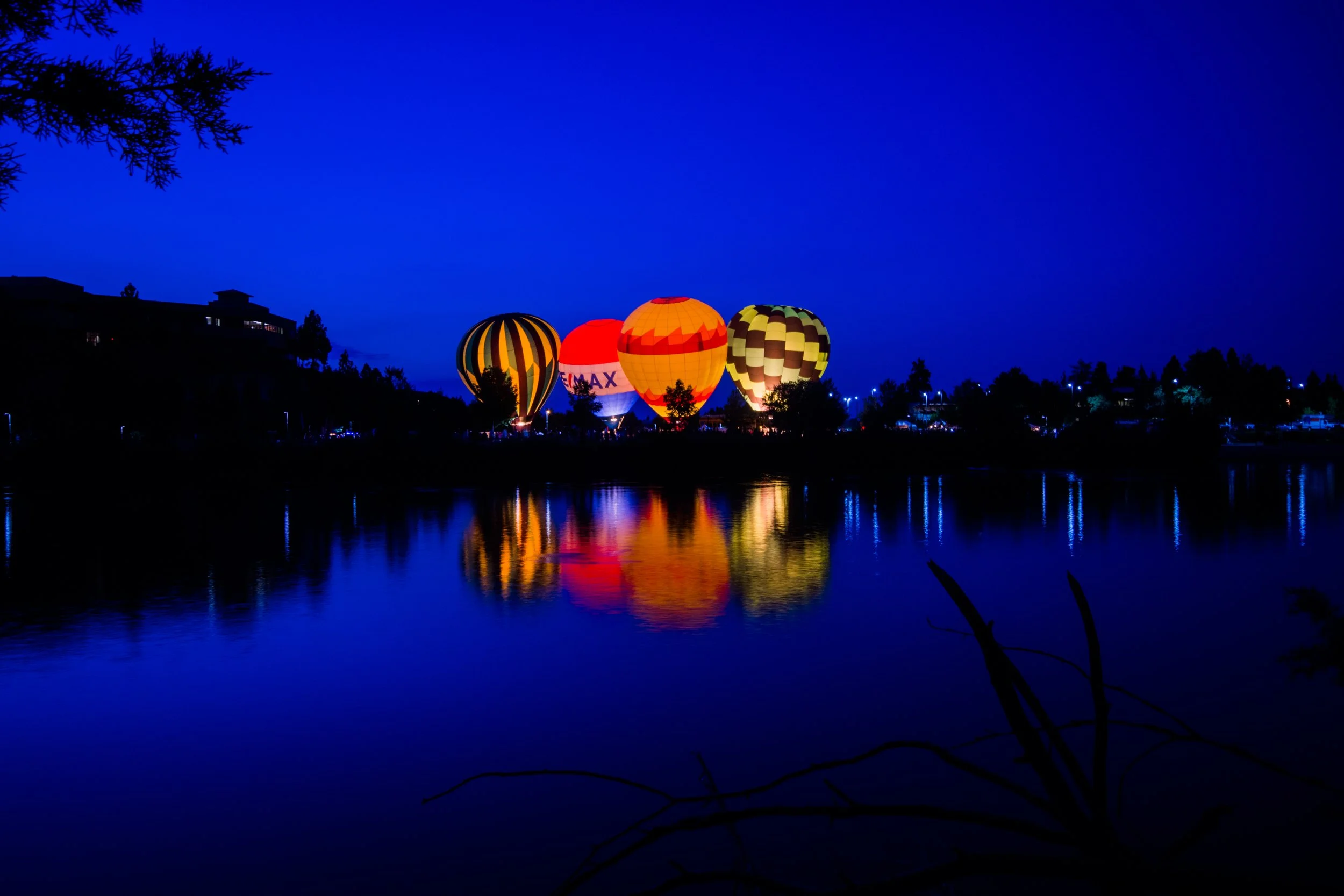 Night scene of illuminated hot air balloons with reflections on a calm lake, set against a deep blue sky.