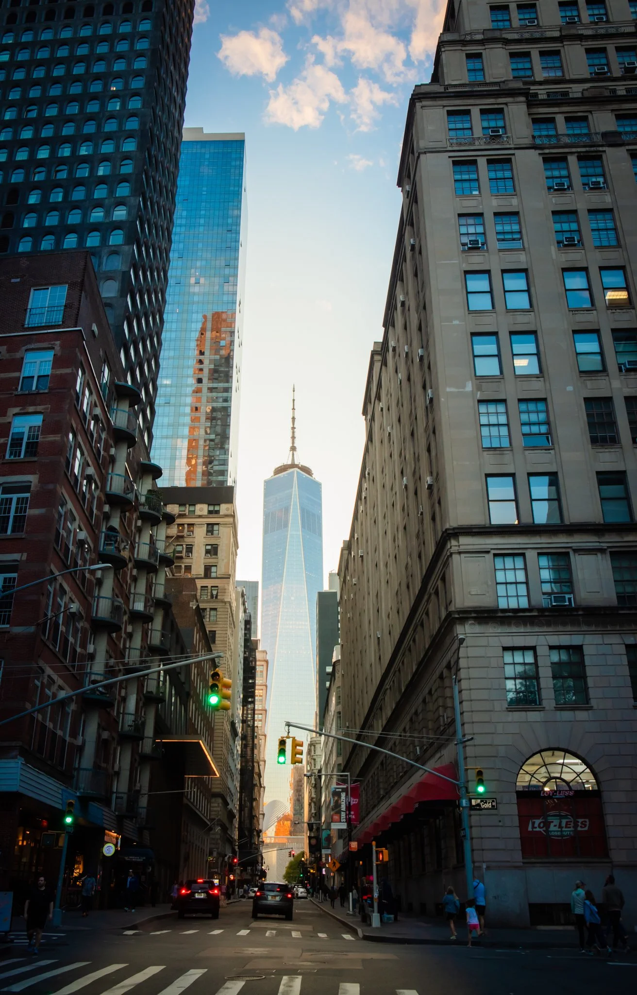 View of a busy city street with tall buildings on either side, featuring the One World Trade Center in the background.