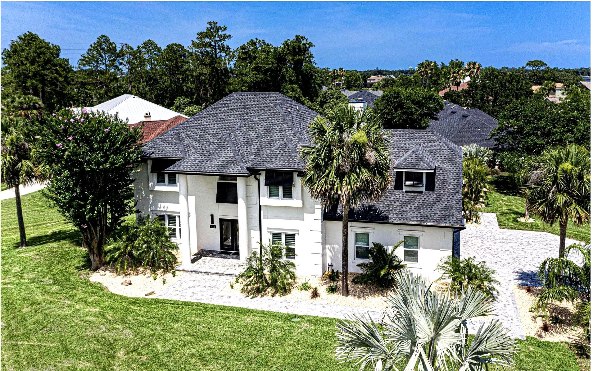 Aerial view of a large white two-story house with a dark gray shingle roof, surrounded by palm trees and a landscaped yard with green grass.