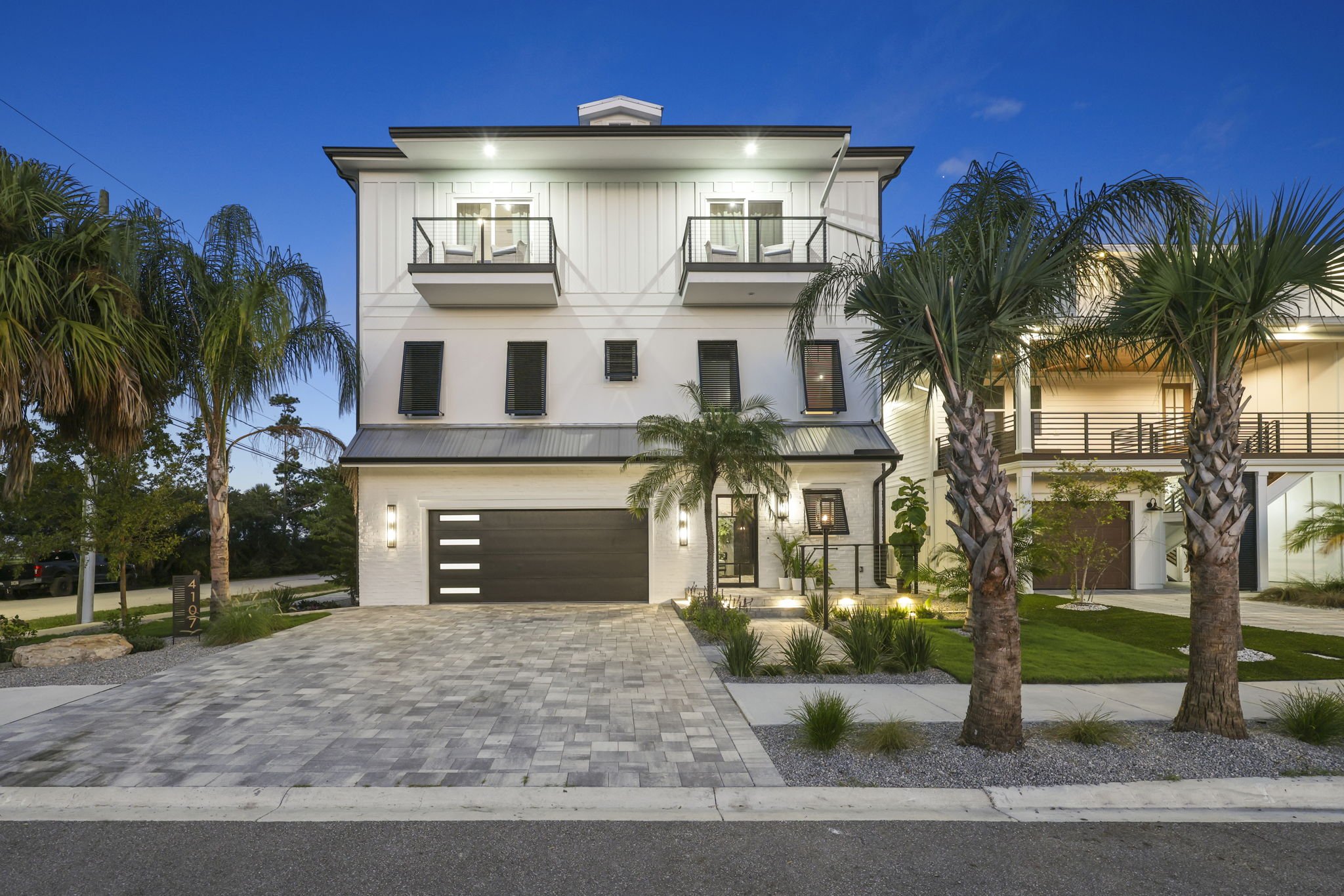 Modern two-story house with a black garage door, two balconies, palm trees, and a paved driveway at dusk.