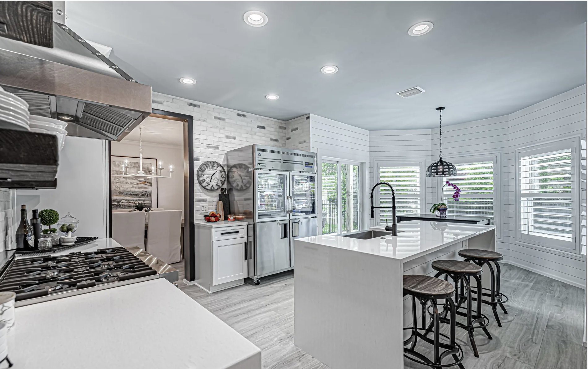 Modern kitchen with white cabinets, stainless steel appliances, large island with bar stools, and dark faucet. There are wall clocks, a flower vase, and a dining area with chairs visible in the background. The room has light wood flooring and large w