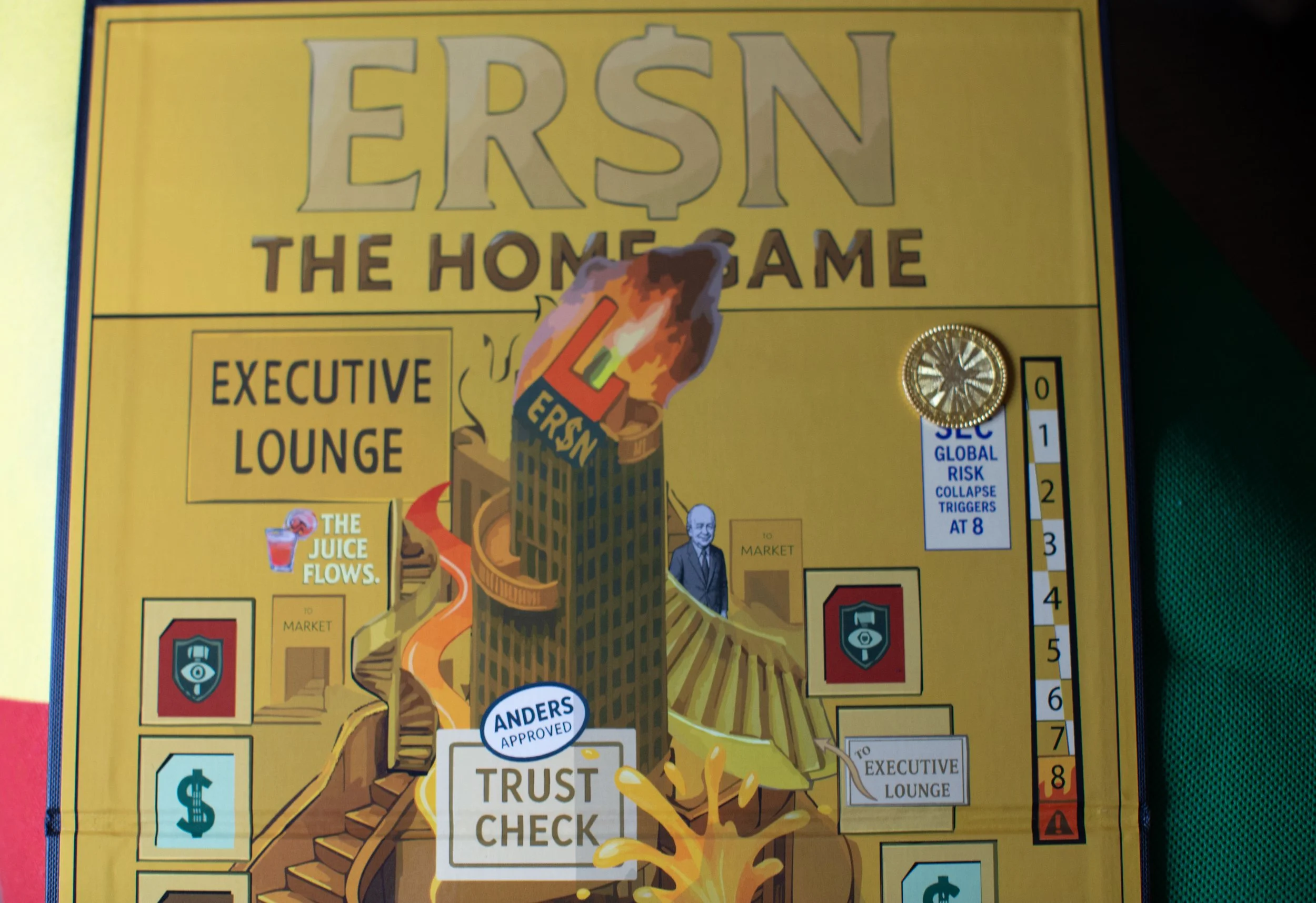 A colorful board game titled "Er$n the Home Game" with a large burning dollar sign on a tall building labeled "Er$n," and pathways leading to areas labeled "Executive Lounge" and "Market." There are various icons, a gold coin, and cards around the board, with some text about risk and trust checks.