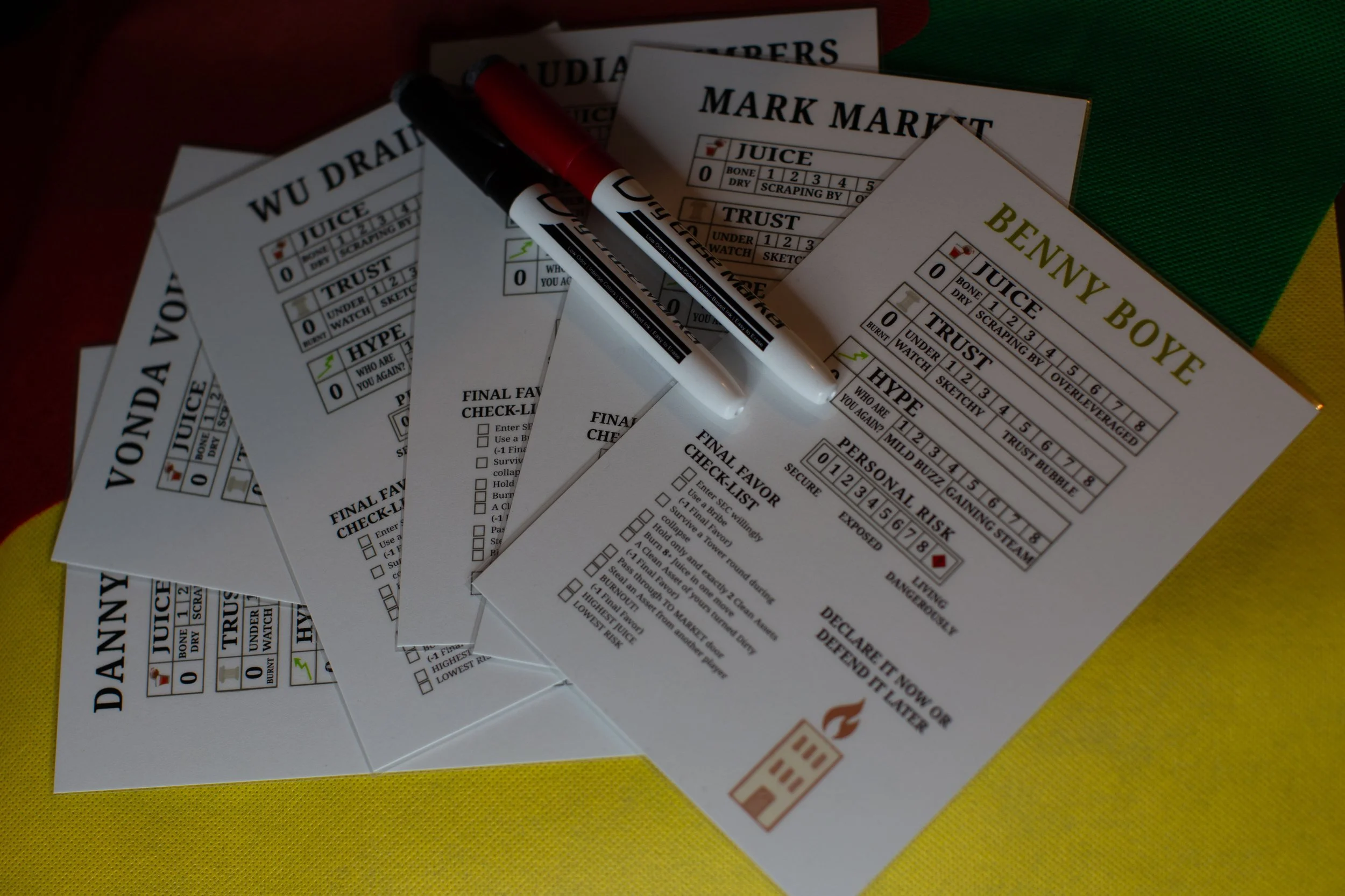 Several voting ballots with checkboxes and markers on top, including ballots labeled 'Vonda Vo,' 'Wu Dra,' 'Mark Mart,' 'Benny Boy,' and others, placed on a yellow surface with a partially visible green and yellow background.