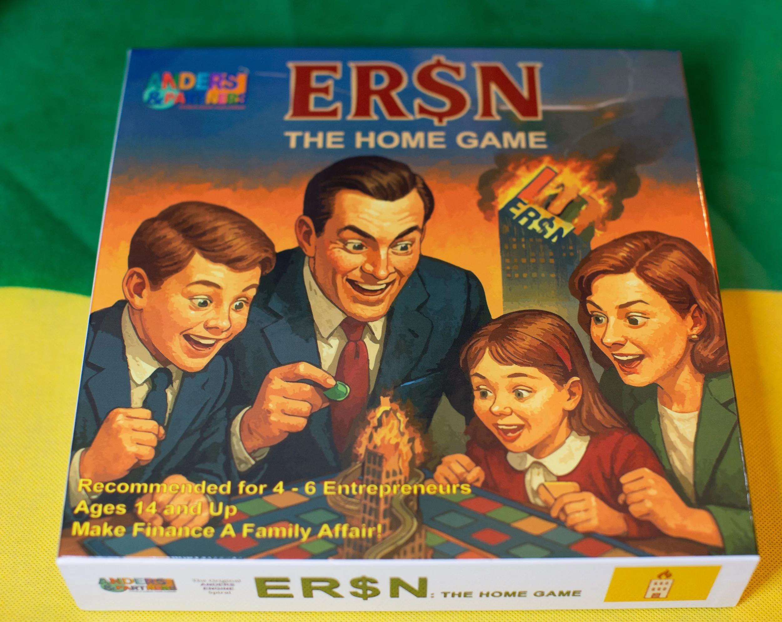 A colorful board game box titled "ER$N: The Home Game" features an illustration of a family of four with expressions of excitement and surprise, playing a game where a small model building is on fire in the center. The background shows a burning skyscraper labeled "ERSN" with smoke billowing from it. The box indicates the game is recommended for 4-6 entrepreneurs, ages 14 and up, with a tagline "Make Finance A Family Affair!" The top left corner has a logo with colorful letters spelling "Anders & Poe."