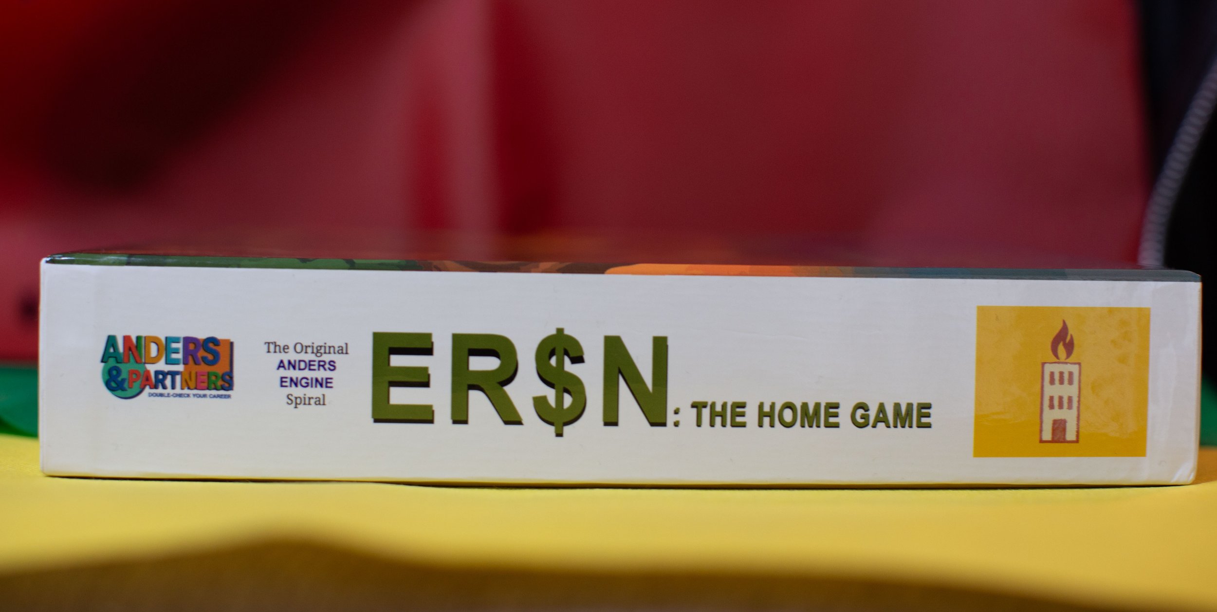 Close-up of a board game box titled 'ERSÑ: THE HOME GAME' with colorful 'ANDERS & PARTNERS' logo on the left and a yellow square with a red candle illustration on the right, placed on a yellow surface.