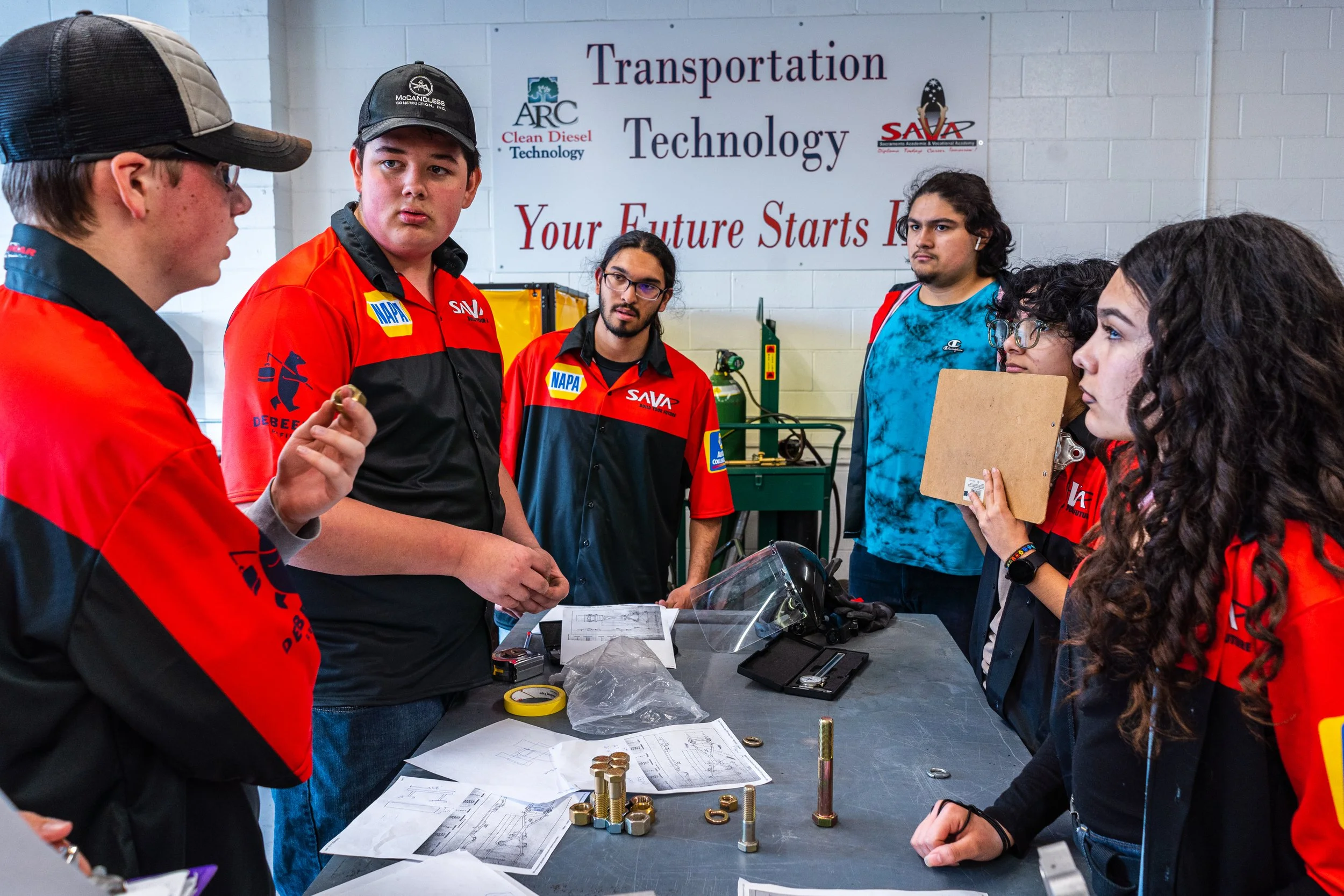 Group of students and instructor in a classroom or workshop setting, engaged in discussion about mechanical parts and engineering projects. There is a large sign in the background that reads 'Transportation Technology' and 'Your Future Starts Here.'