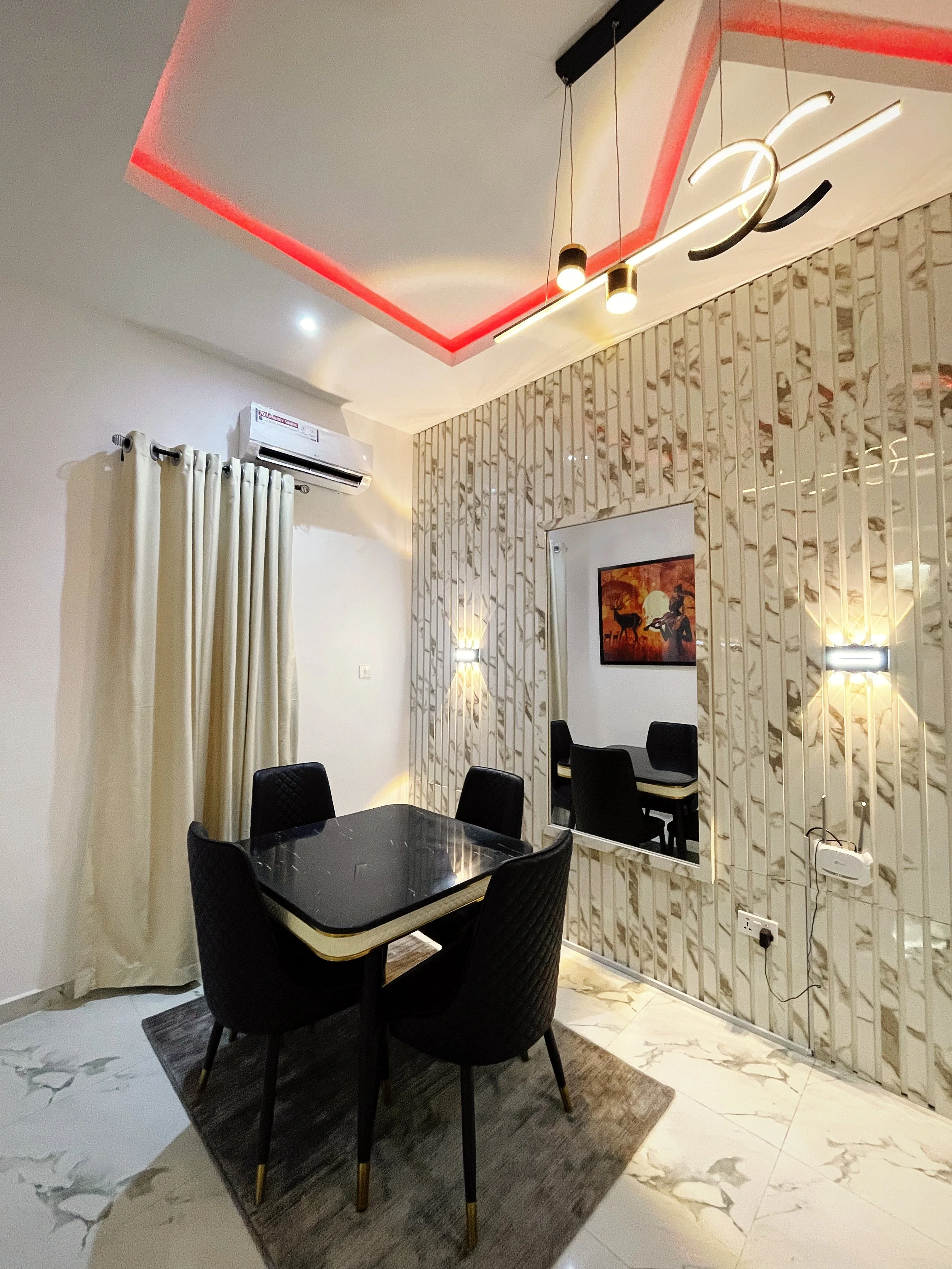 Modern dining room with a black marble table, four black quilted chairs, and stylish lighting under a ceiling with red accents. The room features a decorative wall panel, a large mirror, and beige curtains beside an air conditioning unit.