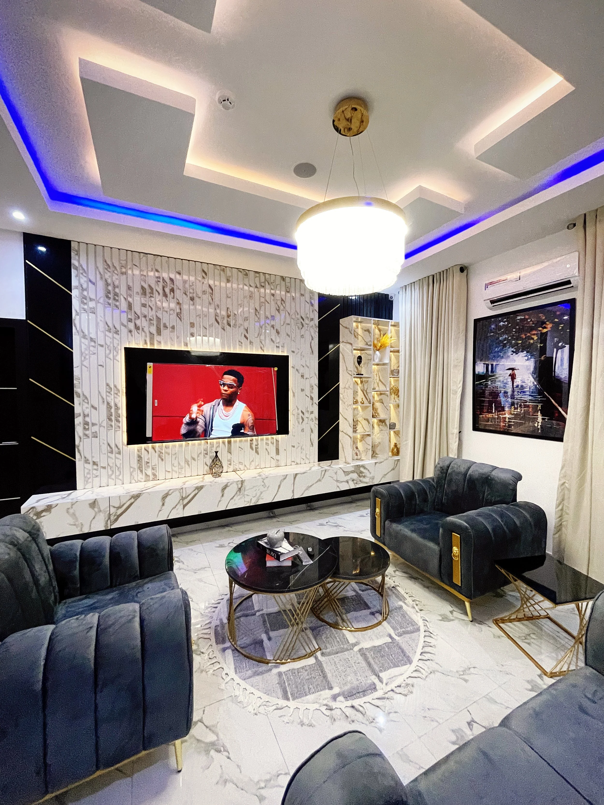 Modern living room with plush grey sofas, a large wall-mounted TV displaying a person, a circular black coffee table with gold accents, white and gold decorative walls, a round pendant light, and abstract wall art. The room has a glossy white tiled floor and cream curtains.