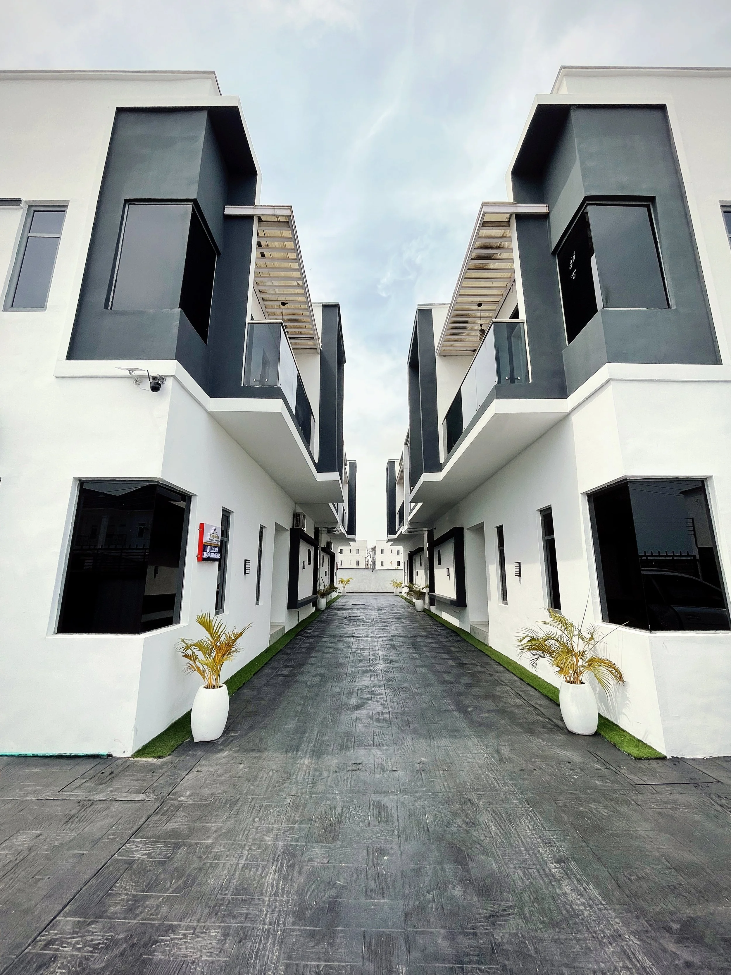 Modern duplex houses with dark gray accents and glass balconies lining a paved driveway, with potted plants placed near the entrances, under a cloudy sky.