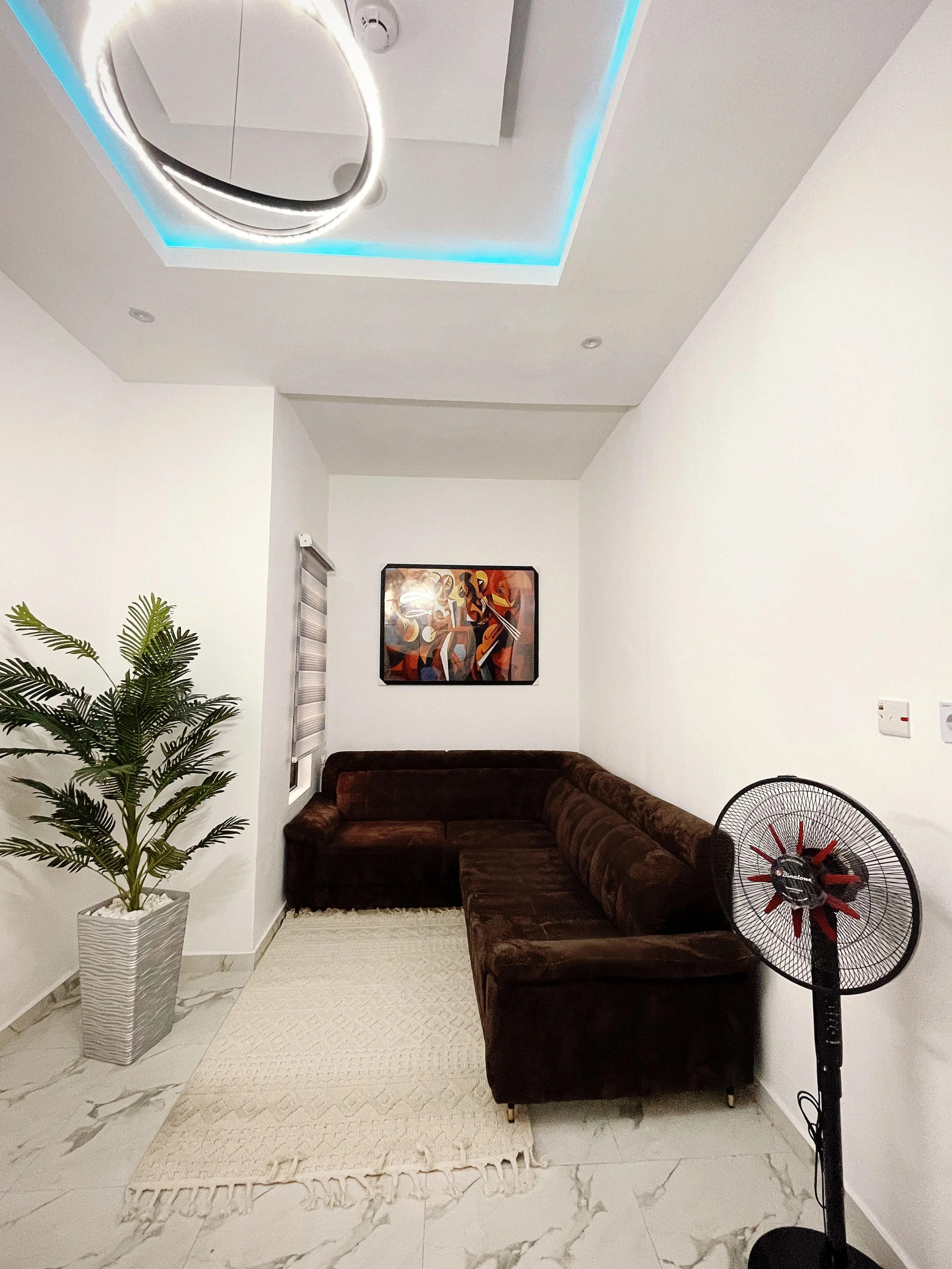 Modern living room with brown L-shaped sofa, abstract painting on the wall, potted plant, white rug, circular ceiling light, and a standing fan.