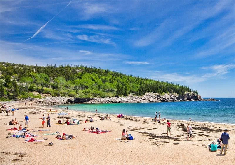 People relaxing and walking on a sandy beach with a green wooded hill and blue sky in the background under a few clouds.
