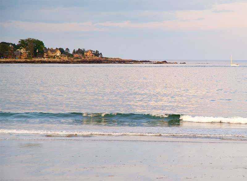 Calm ocean with gentle waves, distant shoreline with houses, and a sailboat.
