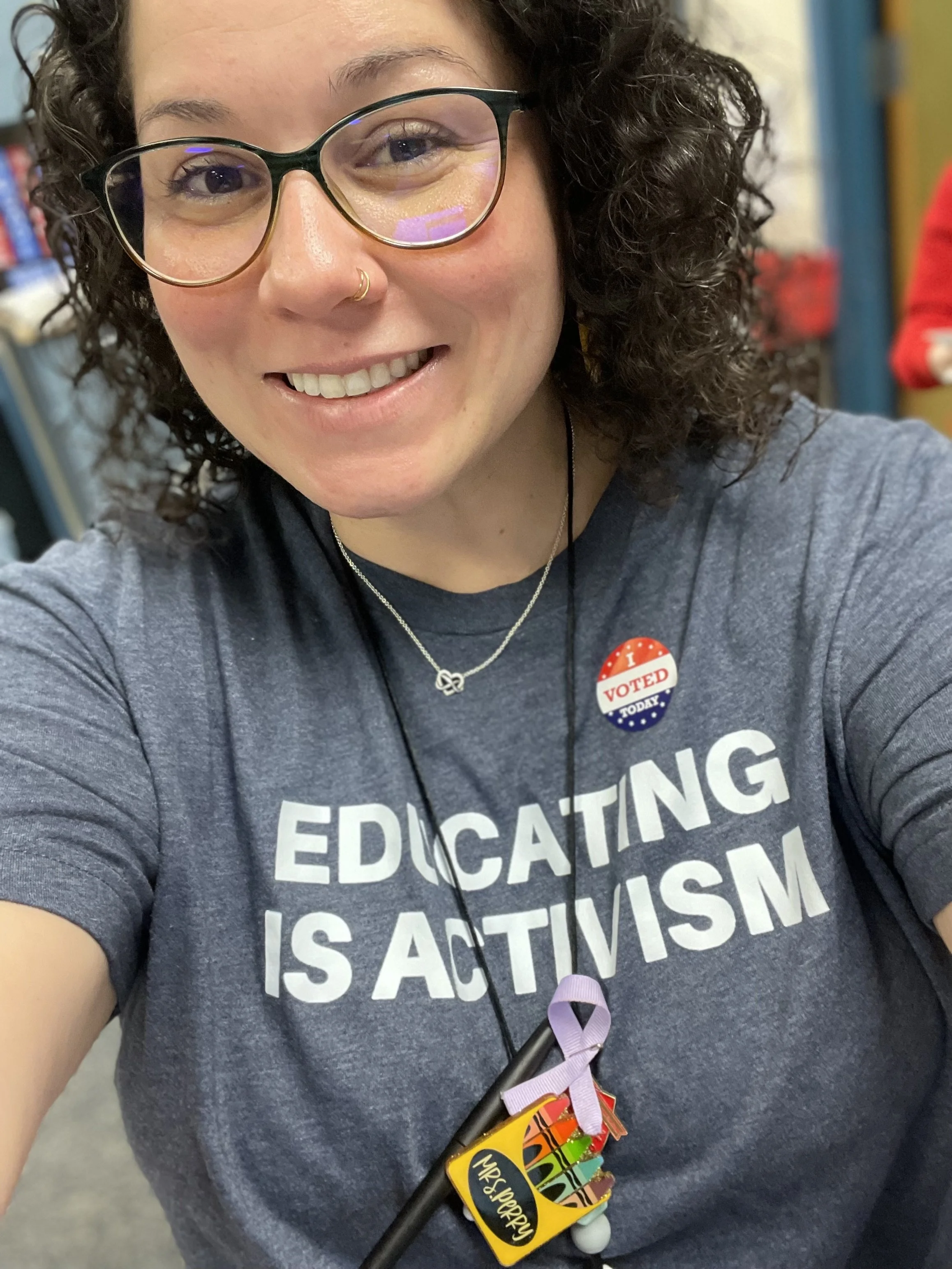 A woman with curly brown hair and glasses smiling; she is wearing a grayish blue shirt with white letters that says Educating Is Activism; she has a lanyard around her neck.