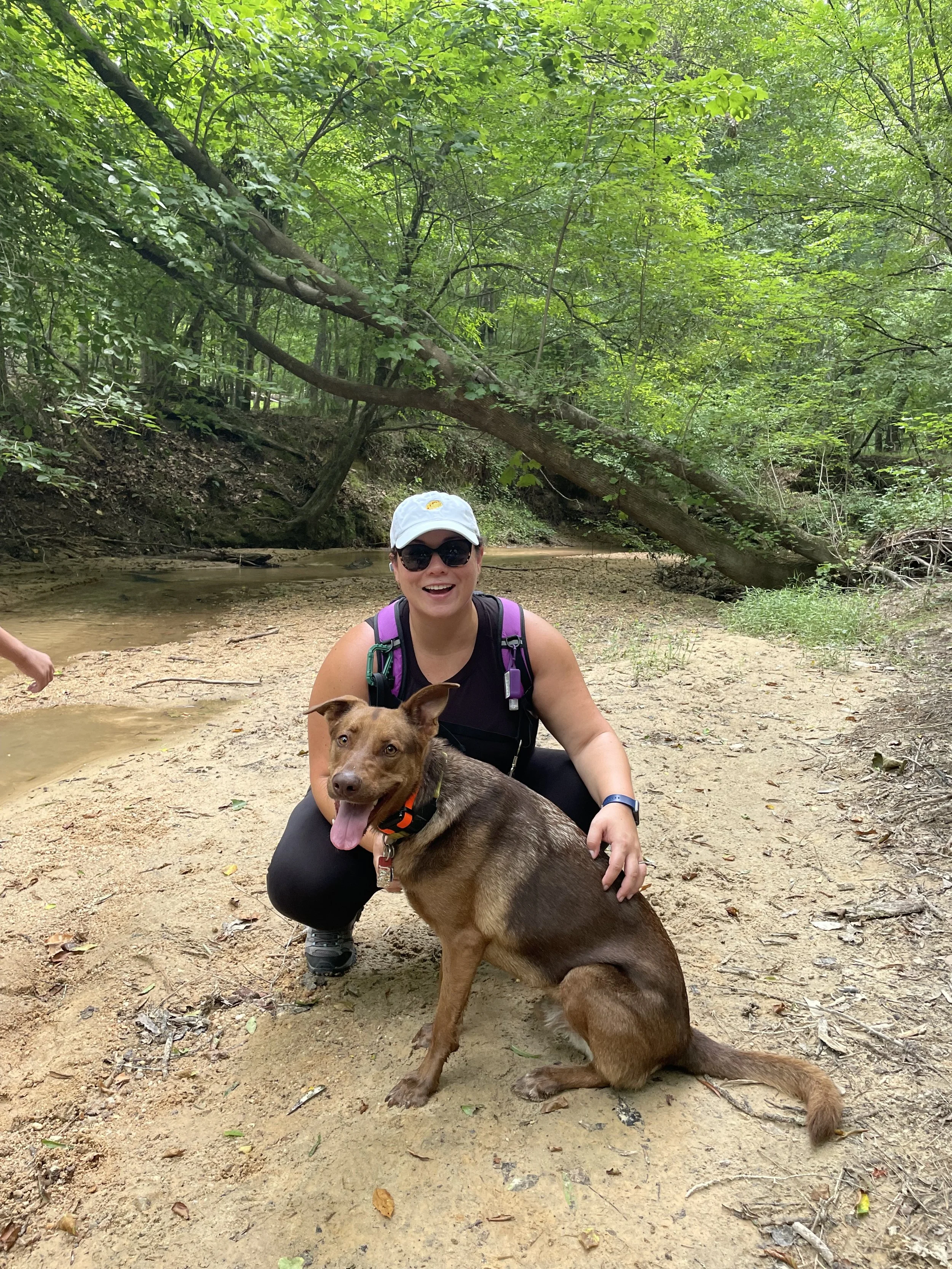 A woman and a brown dog on a sandy patch near a stream; wood are around and behind us. The woman is wearing a hat and sunglasses.