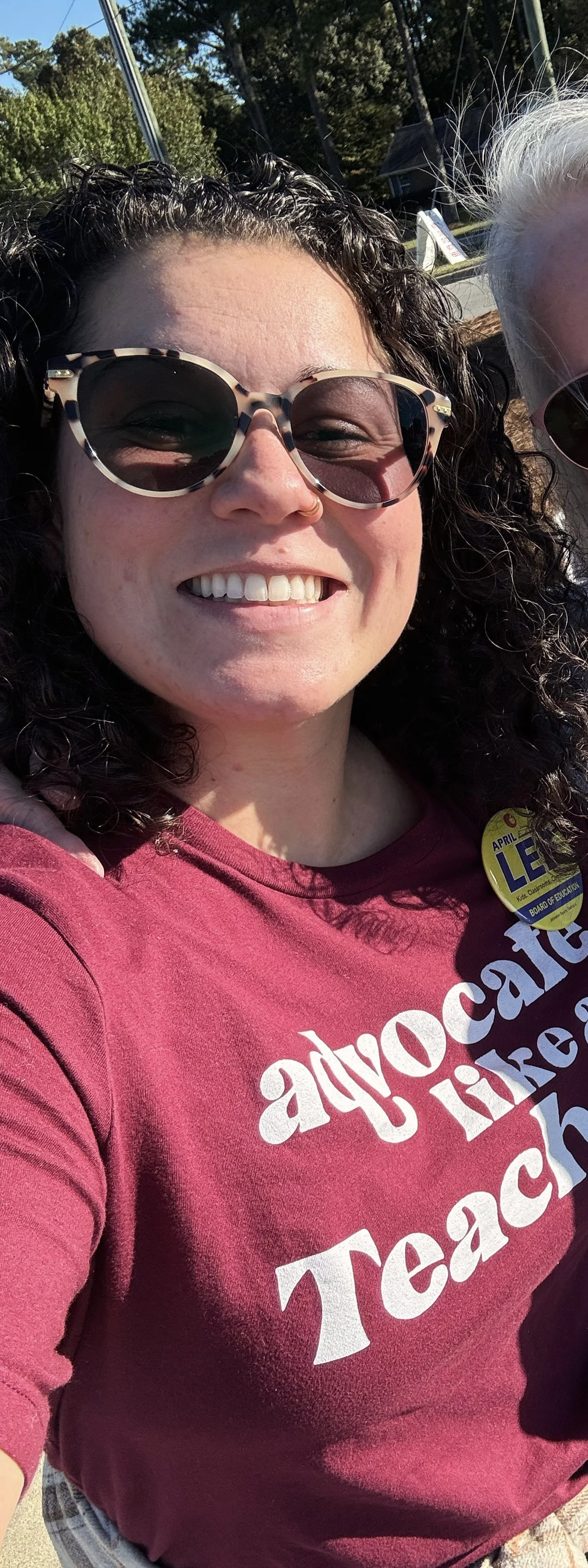 A woman with curly brown hair and glasses smiling; she is wearing a maroon shirt that says Advocate Like A Teacher