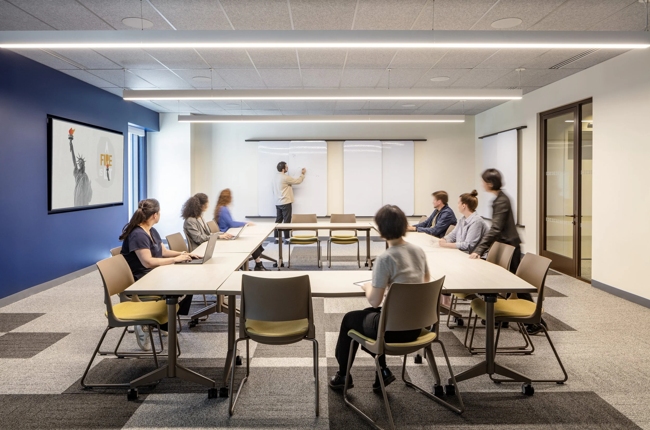 A business meeting in a modern conference room with people sitting around a U-shaped table, some using laptops, while a man at the front writes on whiteboards.