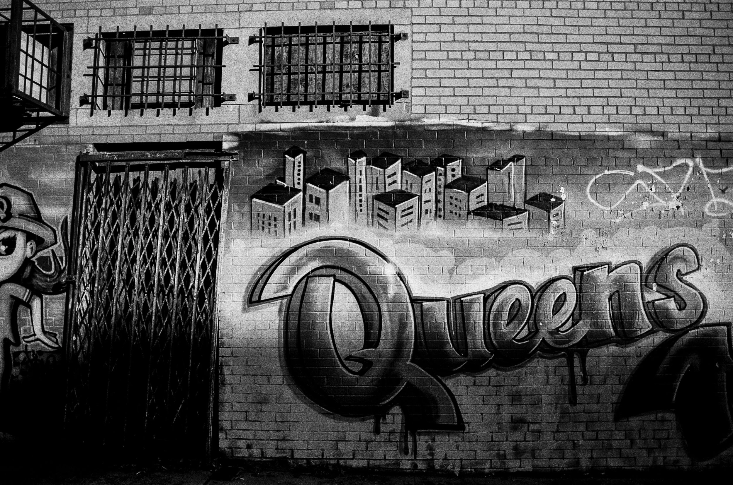 Black and white graffiti mural on a brick wall featuring the word "Queens" in large, stylized letters with a cityscape of buildings above it and a cartoon character wearing a hat on the left side.