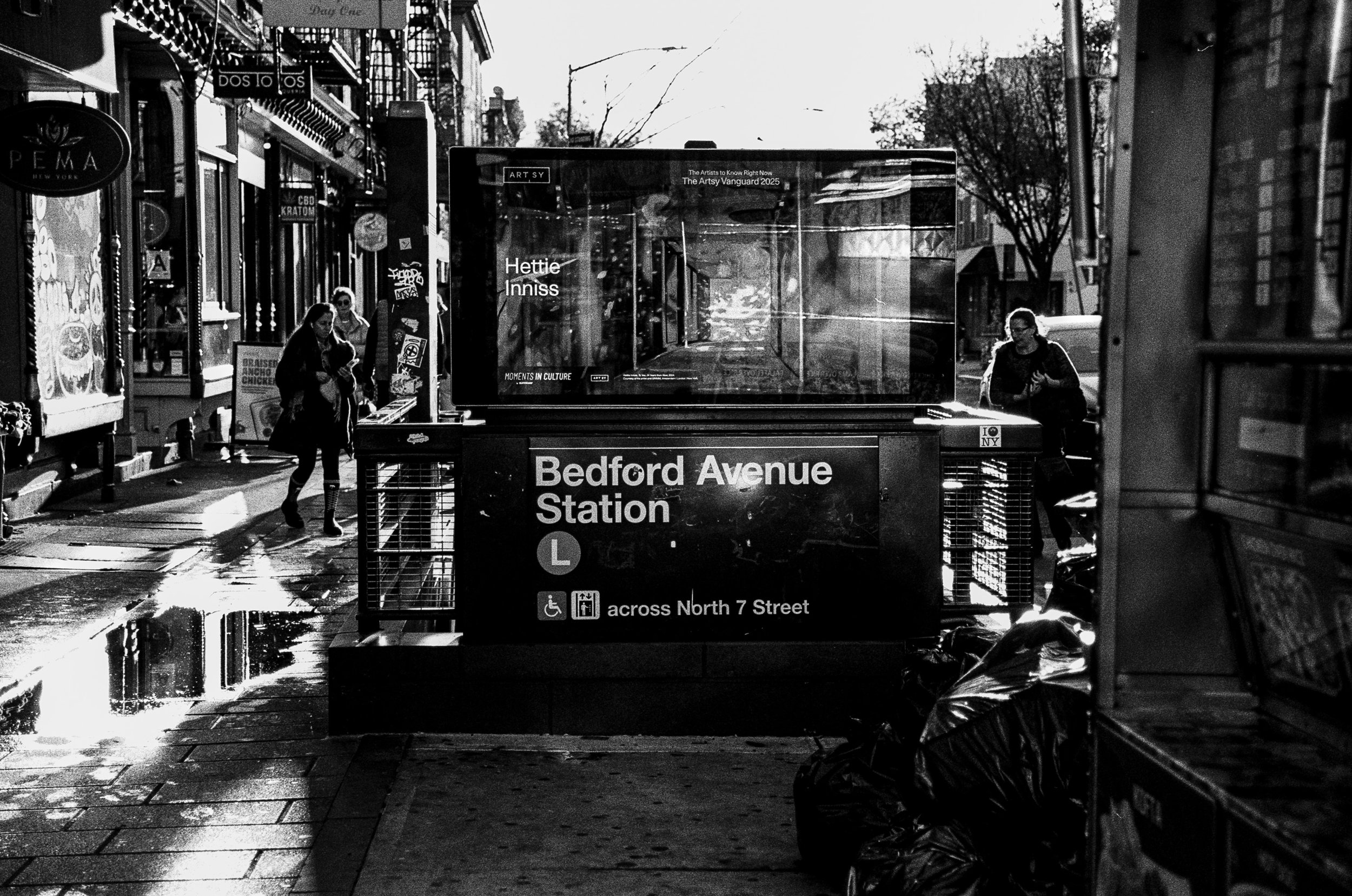 Black and white photo of Bedford Avenue subway entrance in Brooklyn, New York. The sign indicates it is near North 7 Street, and there are a few pedestrians on the street with storefronts and a wet sidewalk visible.