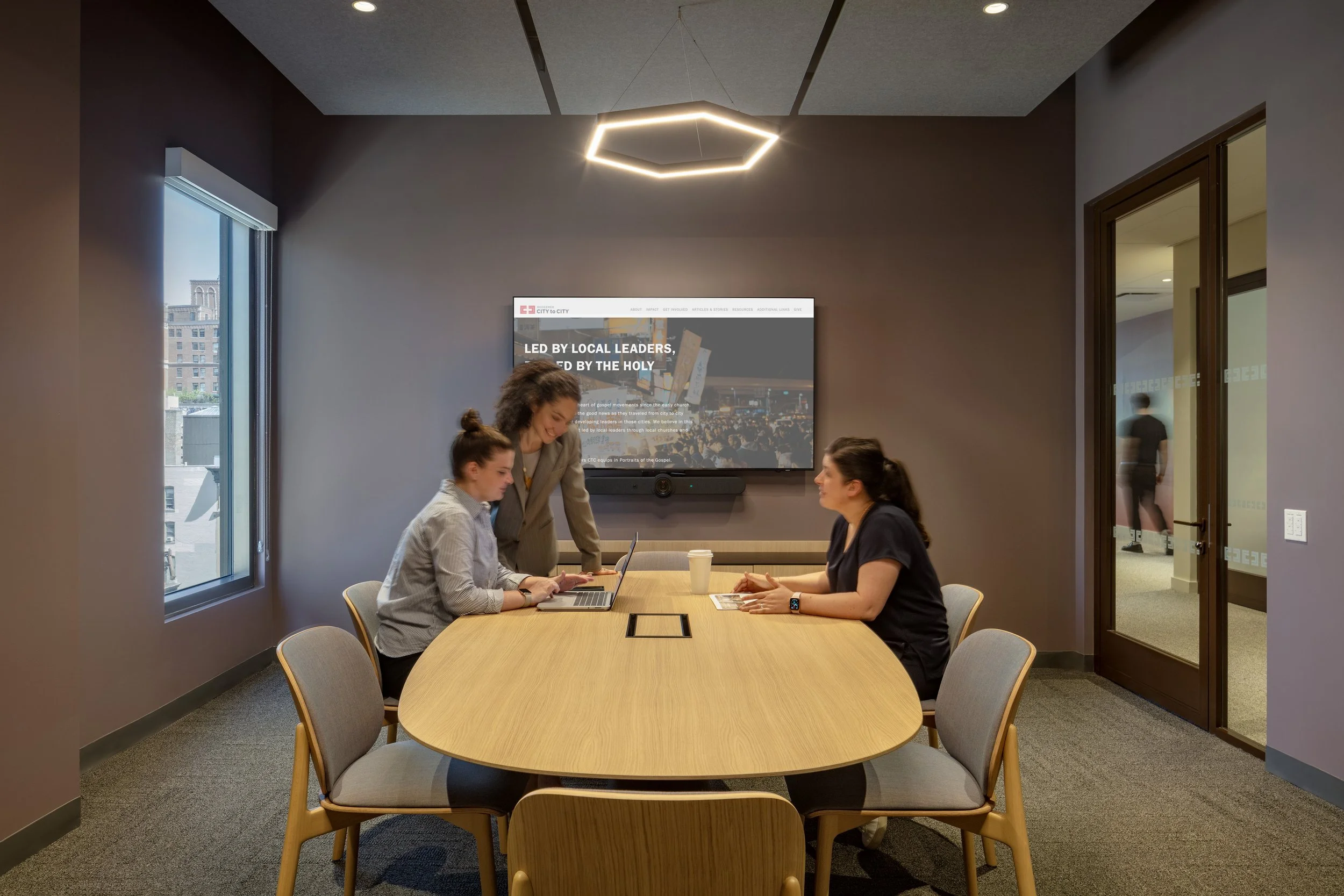 Three women are in a conference room. Two women are seated at a wooden table, one working on a laptop, and one-looking at a smartphone, while the third woman stands between them, leaning over and talking. The room has a large window, a wall-mounted s