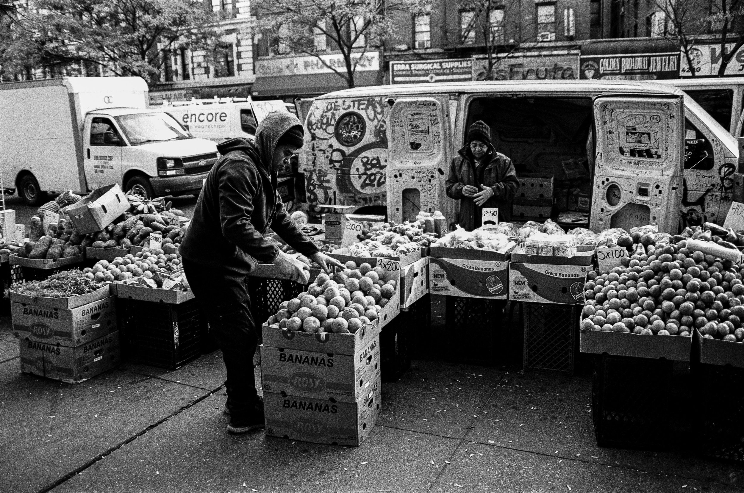 Street scene with vendors selling bananas and other produce from cardboard boxes and a parked van in the background.