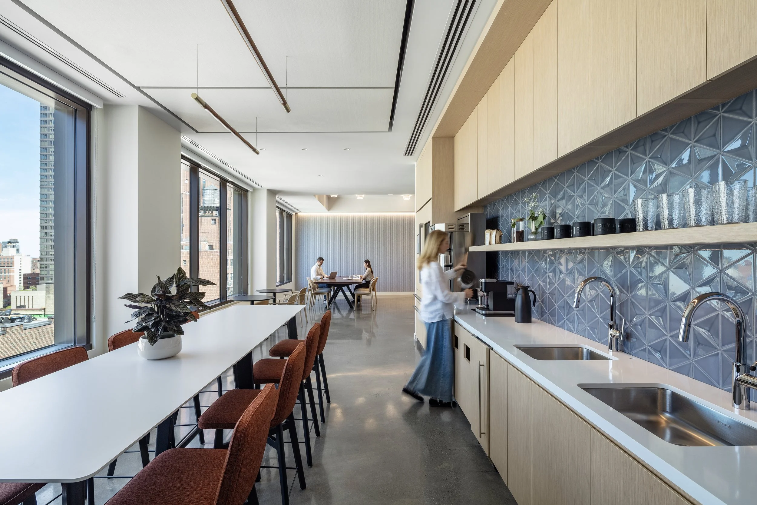 Modern office kitchen and dining area with large windows, a long white table with orange chairs, a plant in a white pot, and a woman preparing coffee. In the background, two people work on laptops at a table.