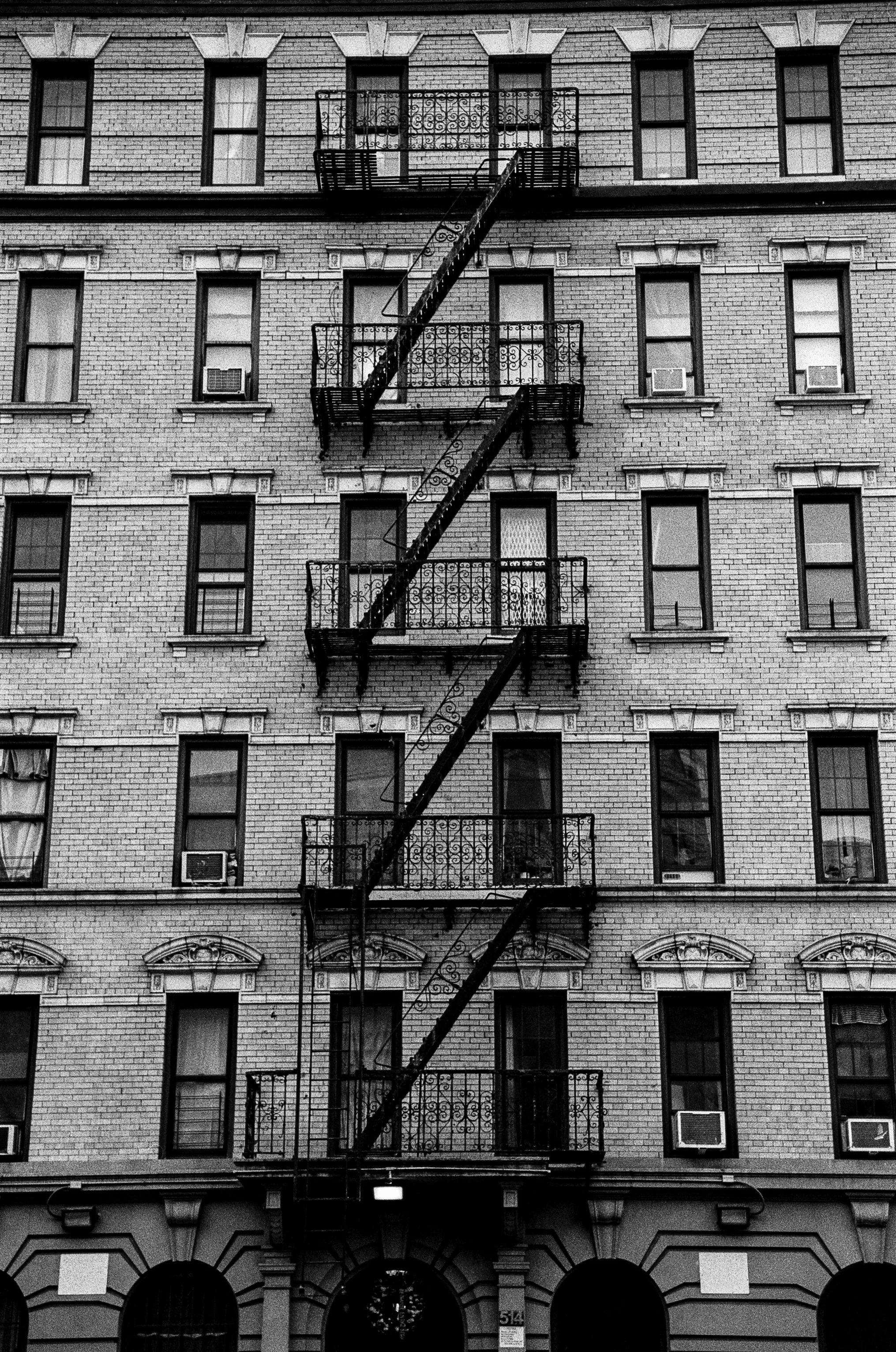Black and white photo of an urban brick building with fire escapes and multiple windows, some with air conditioning units.