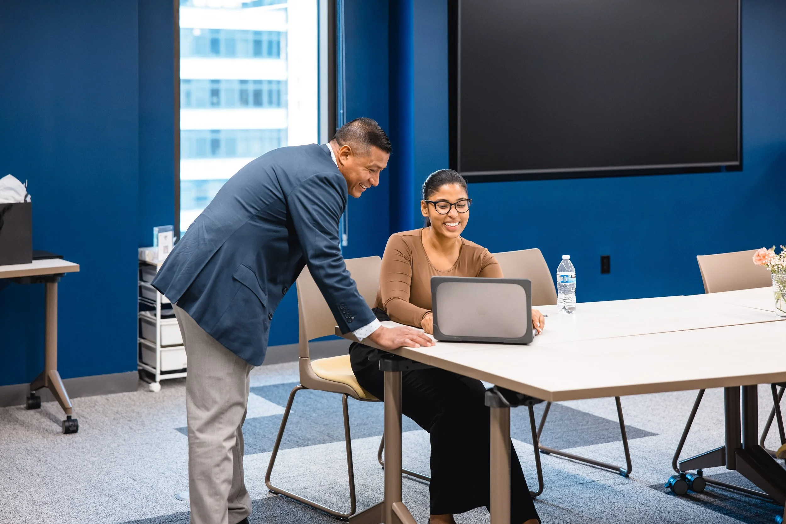 A man in a suit leaning over to talk to a woman at a conference table. The woman is sitting with a laptop open in front of her, smiling, wearing glasses and a brown top. The room has blue walls and large windows, with a water bottle and flowers on the table.