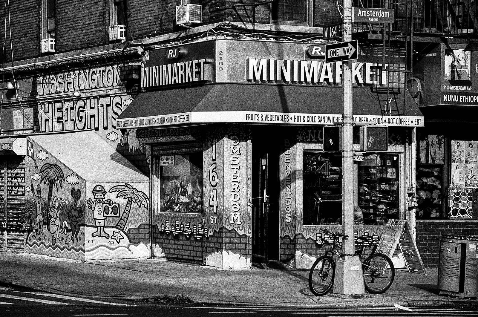 A black and white photo of a corner mini-mart called Washington Heights Mini Market, with street signs for Amsterdam Avenue and W 164 Street. The storefront features colorful murals, advertisements for fruits, vegetables, hot and cold sandwiches, and