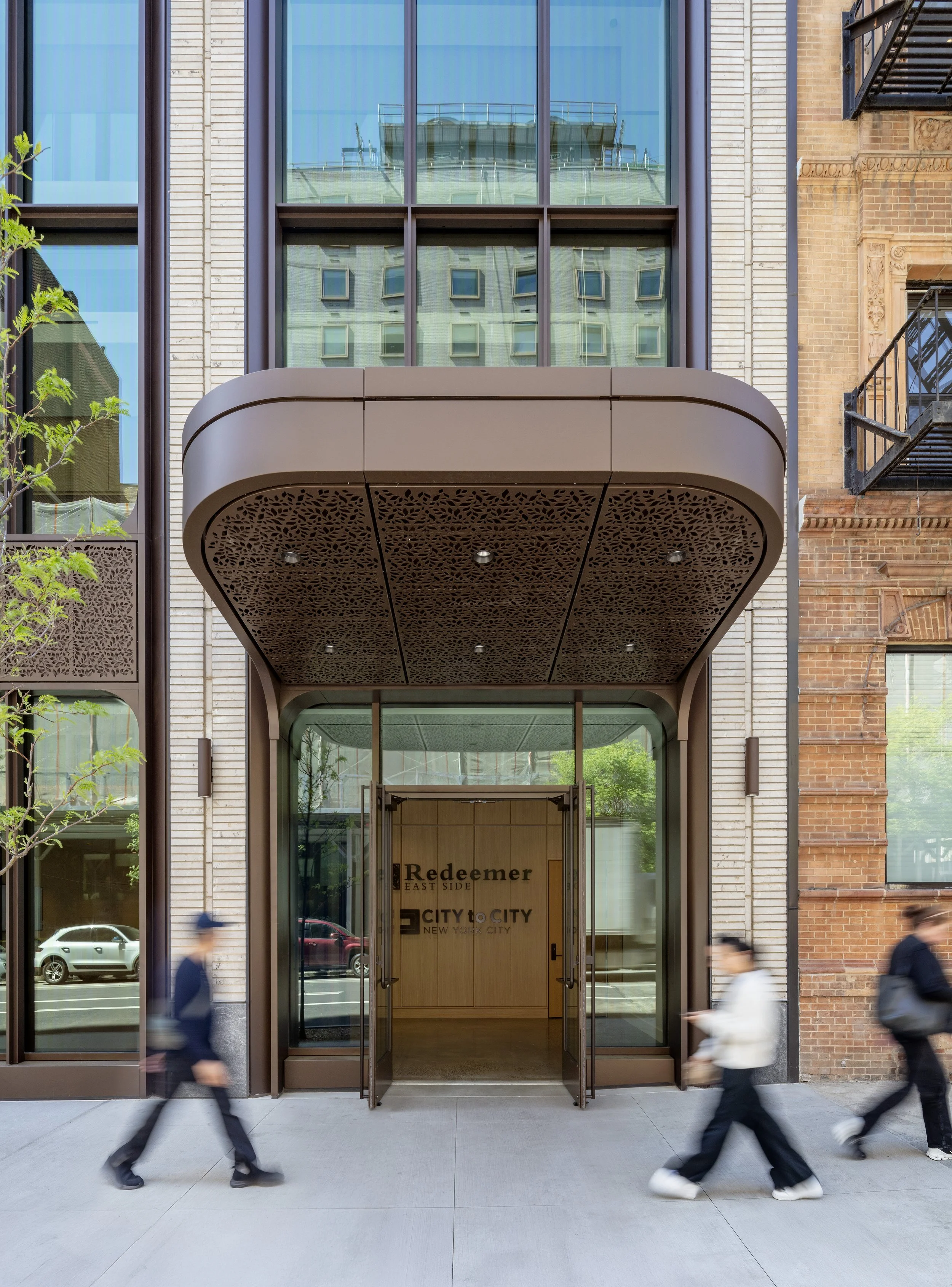 Modern building entrance with glass doors, an overhang with decorative lattice, and pedestrians walking by.