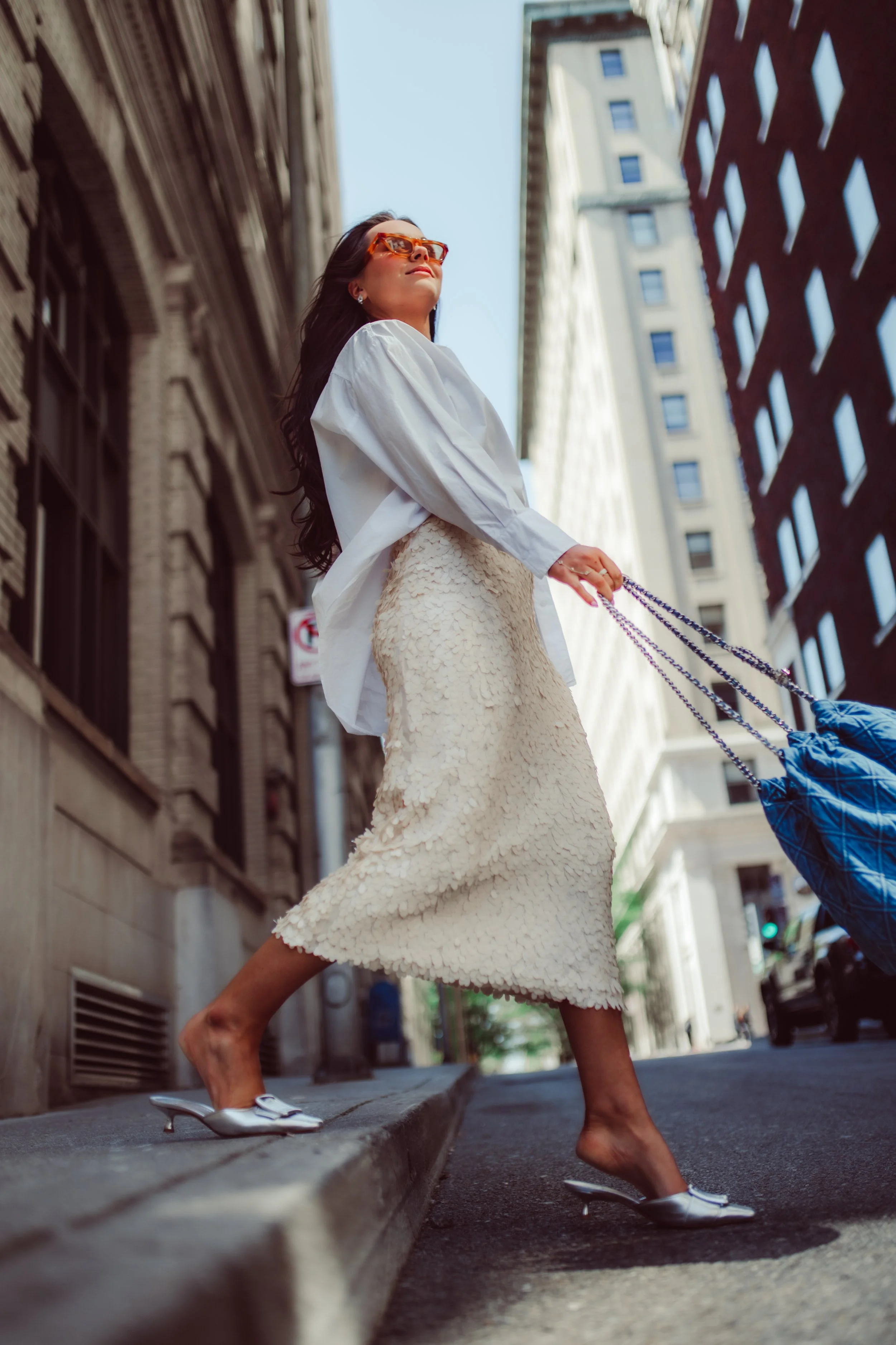 Fashionable woman walking city street wearing sunglasses, white blouse, and textured ivory skirt, carrying a large blue bag.