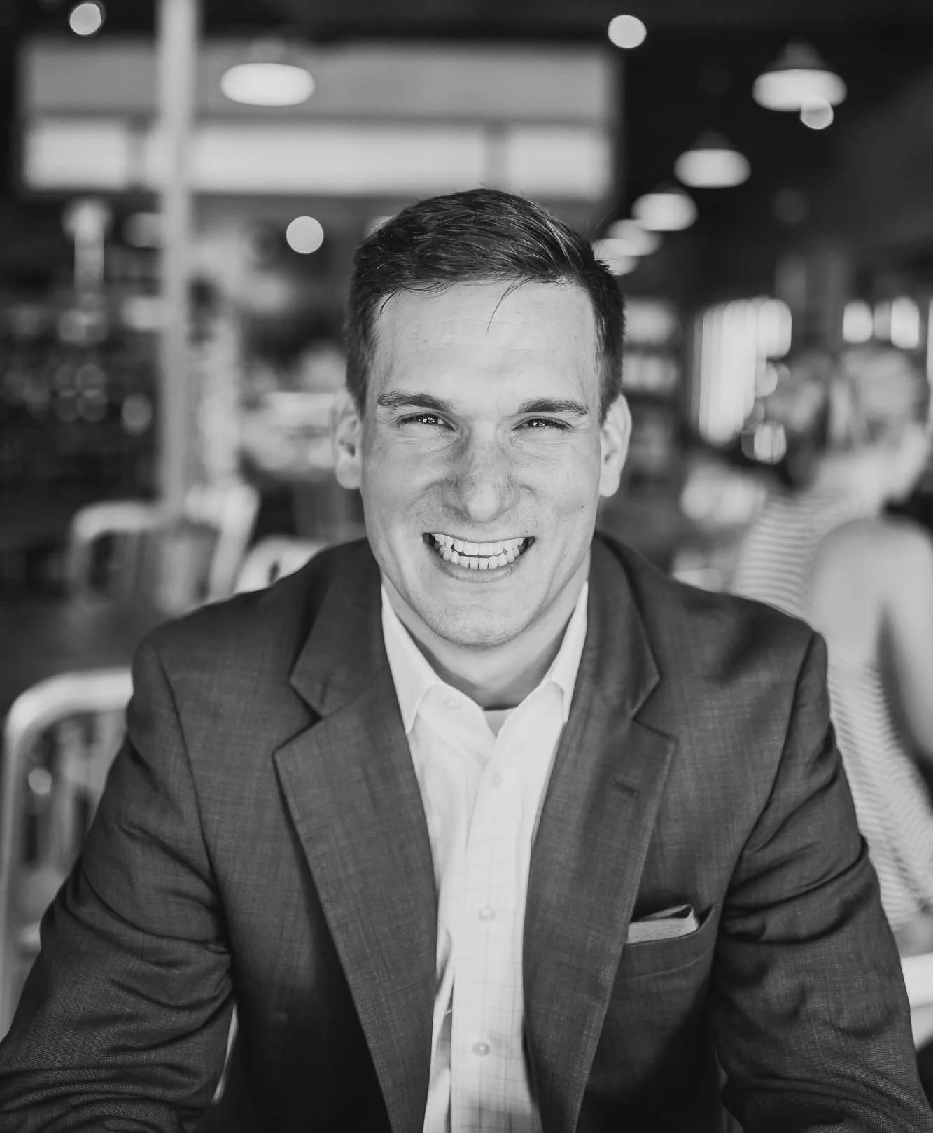 A black and white photo of a smiling man in a suit sitting at a table in a busy restaurant or cafe.