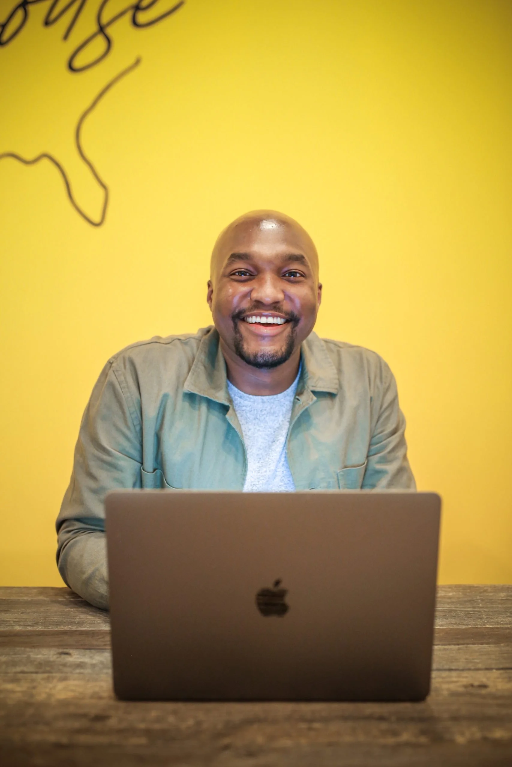 A man smiling while sitting at a wooden table with a closed laptop in front of him, against a yellow wall background.