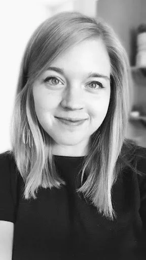 Black and white portrait of a young woman with shoulder-length hair, smiling slightly, wearing a dark top, with a blurred background.