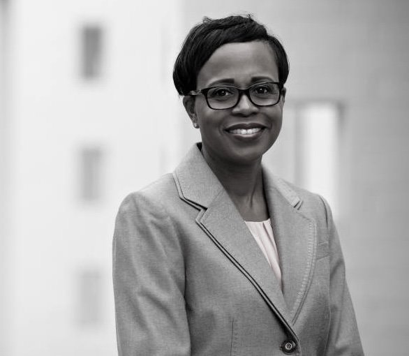 Portrait of a smiling woman with short black hair, glasses, wearing a light-colored blazer, standing outdoors in front of a blurred building.